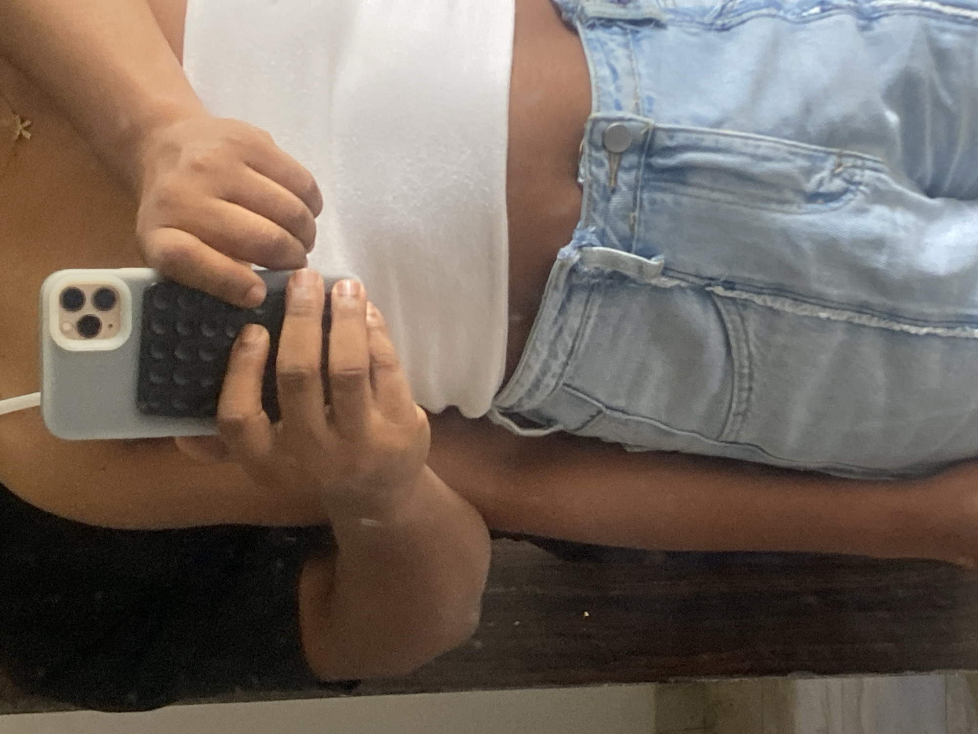 The image shows a person taking a selfie in a mirror. The person is wearing a white tank top and light blue denim overalls. They are holding a grey iPhone with three camera lenses. The background is blurry, but appears to be a bathroom setting. There is no information in the image to identify the location in Abuja, Nigeria, or to confirm the image was taken there at all.