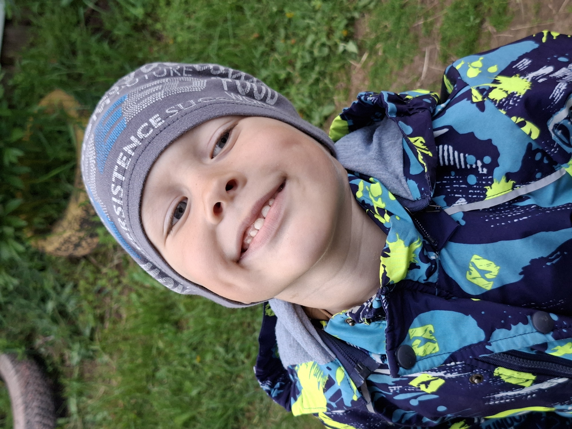 A child is facing the camera with a smile, positioned slightly off-center. The child is wearing a gray beanie with blue accents and patterned text. The jacket is a mix of blue and navy with bright yellow and white designs. The background consists of green grass and hints of dirt or soil. The location and city are Unknown.