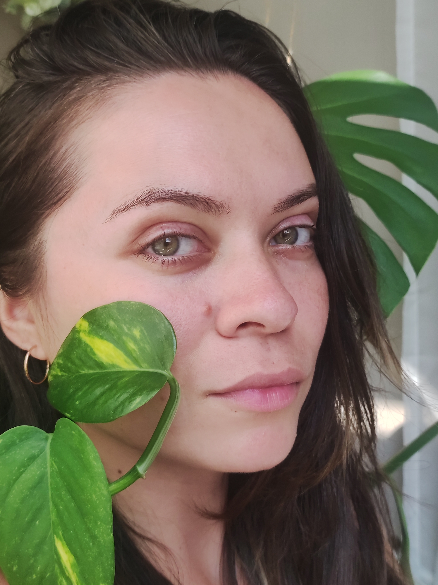 A close-up shot shows the right side of a woman's face, partially obscured by green leaves of a houseplant. She has brown hair and green eyes. The background is out of focus and appears to be a room with a window and natural light. The scene is indoors, likely in a residential setting. There is no information to indicate the location in Chaikovsky, Russia.