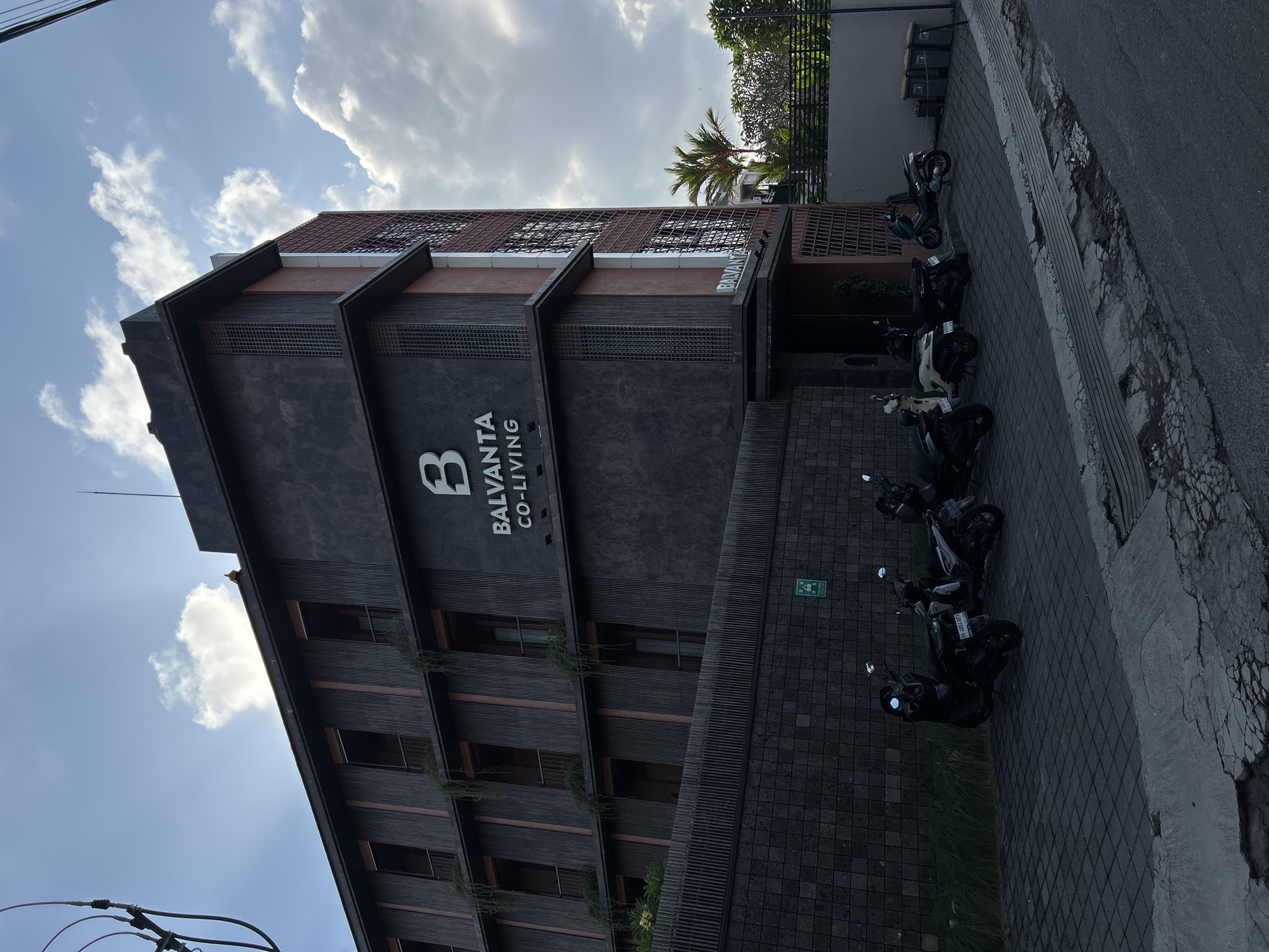 In the city of North Kuta, Indonesia, a multi-story building identified by the signage "BALVANTA CO-LIVING" stands under a partly cloudy sky. The building's exterior features a dark, textured facade with vertical wooden slats and balconies. Several motorbikes are parked on a paved area in front of the building. To the right of the building, a palm tree and lush green foliage are visible behind a wall. A paved street runs along the foreground, showing some wear and tear. The overall scene depicts an urban environment with residential and commercial elements.