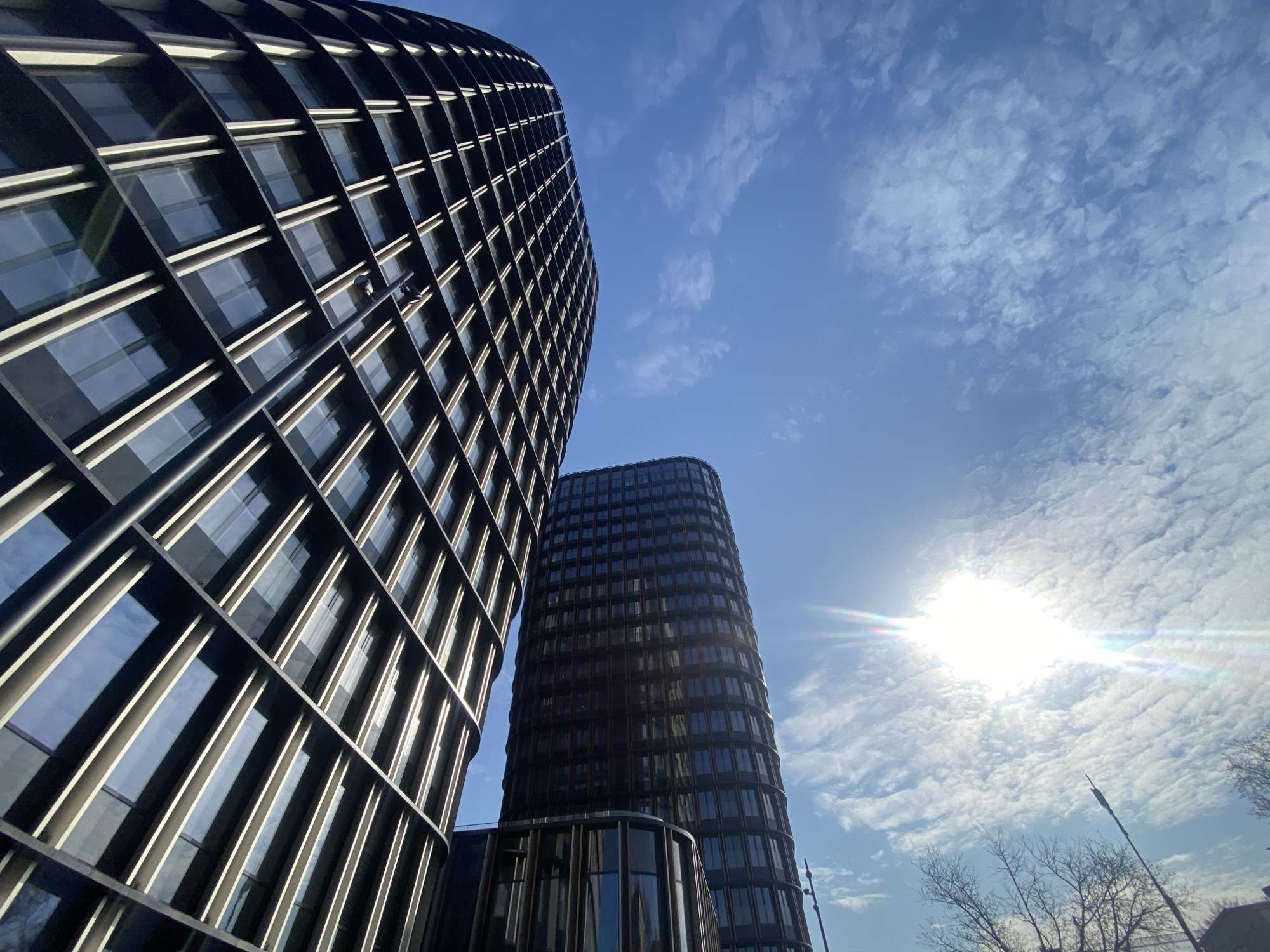 The image depicts a low-angle view of two modern, curved, dark-colored high-rise buildings in Moscow, Russia, set against a bright blue sky with scattered clouds and a strong sun. The building on the left is taller and shows a grid-like facade with alternating dark and light segments, presumably windows and support structures. The building on the right is shorter and has a more cylindrical shape. Both buildings feature numerous windows. The lower part of the right building transitions into a smaller, rounded structure with glass panels. Trees with bare branches are visible in the lower right corner, suggesting it might be late fall or winter. A tall lamppost is partially visible in the bottom right of the frame. The sun shines brightly behind the shorter building, creating lens flare. The sky contains patchy clouds.