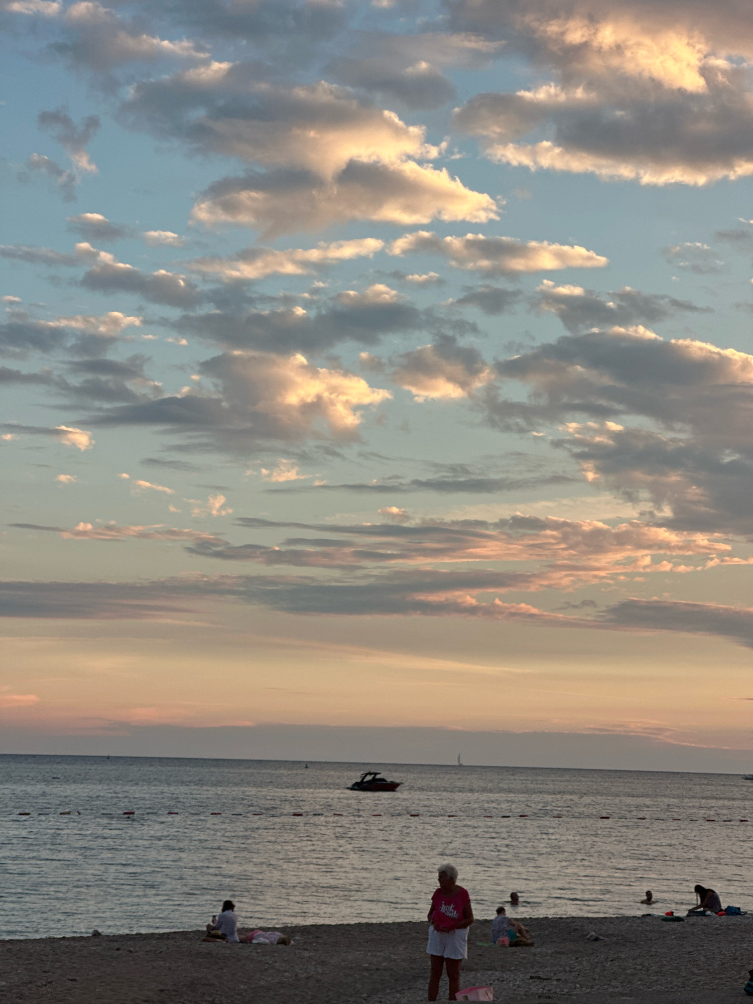 The image depicts a serene beach scene in Budva, Montenegro, at dusk. The sky is a canvas of soft blues and pinks, with scattered clouds catching the warm glow of the setting sun. A calm sea stretches to the horizon, where a small boat and a sailboat are visible in the distance.
On the pebbled beach, several people are enjoying the evening. A woman in a bright pink t-shirt and white shorts stands prominently in the foreground, holding a circular object. Further along the beach, other individuals are relaxing, with some seated and others wading in the shallow water. The scene is framed by the natural beauty of the coastline, with the gentle waves lapping at the shore.