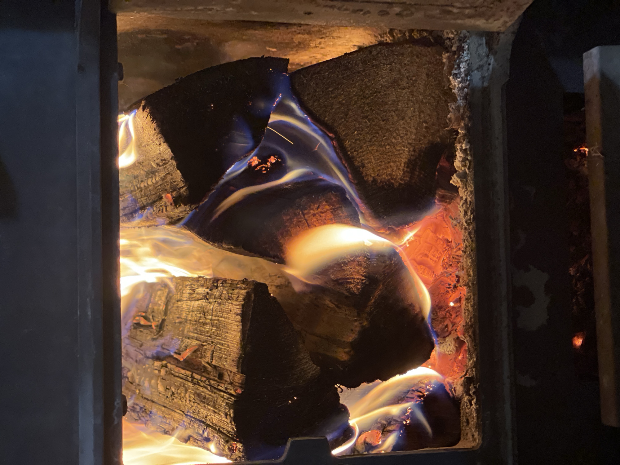 The image shows a close-up view of burning wood inside a wood-burning stove or fireplace. The wood logs are arranged in a stack and are actively burning, producing flames of various colors, including yellow, orange, and blue. The interior walls of the stove appear to be made of a textured, possibly fire-resistant material. The scene is contained within a dark, enclosed space, likely the firebox of the stove. No people are visible in the image. There is no indication this is in the city of Перевозное, Russia.