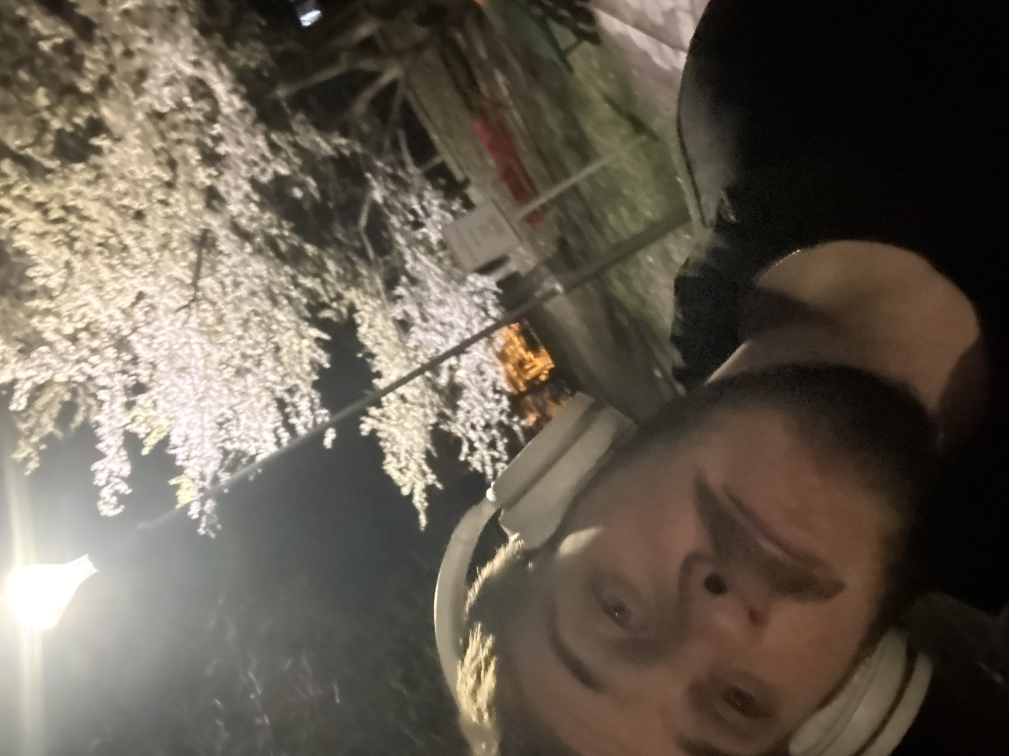 A selfie of a man with headphones at night. The background features illuminated trees, likely decorative lighting. The location is potentially in Sofia, Bulgaria.