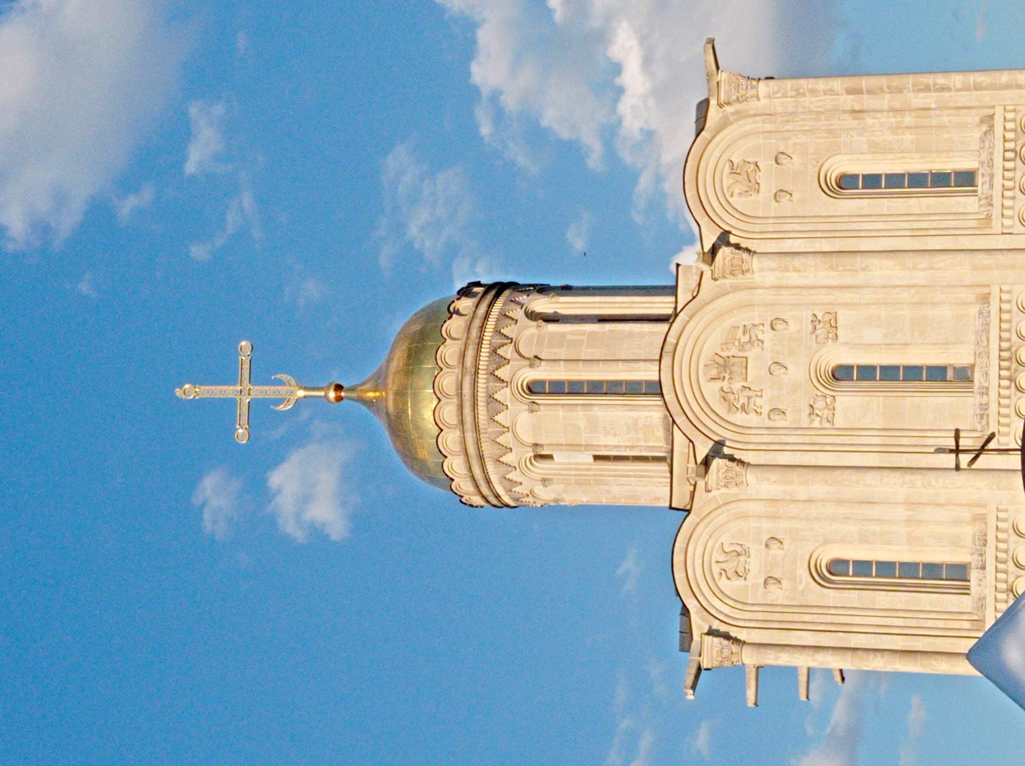 This is a vertically oriented, close-up, outdoor, eye-level shot of a portion of a building's facade, featuring a golden dome topped with a cross. The building appears to be made of light-colored stone and displays intricate architectural details, including arched windows, decorative carvings of animals and geometric patterns, and columnar elements. The sky is a vibrant blue with scattered white clouds, suggesting a clear day. The lighting indicates it is daytime, likely late morning or early afternoon, casting soft shadows that highlight the building's textures. The perspective from below looking up emphasizes the height and grandeur of the structure. There is no visible text in the image. The image's orientation and content suggest a religious or historical building, possibly a church or cathedral. No people or explicit activities are depicted.