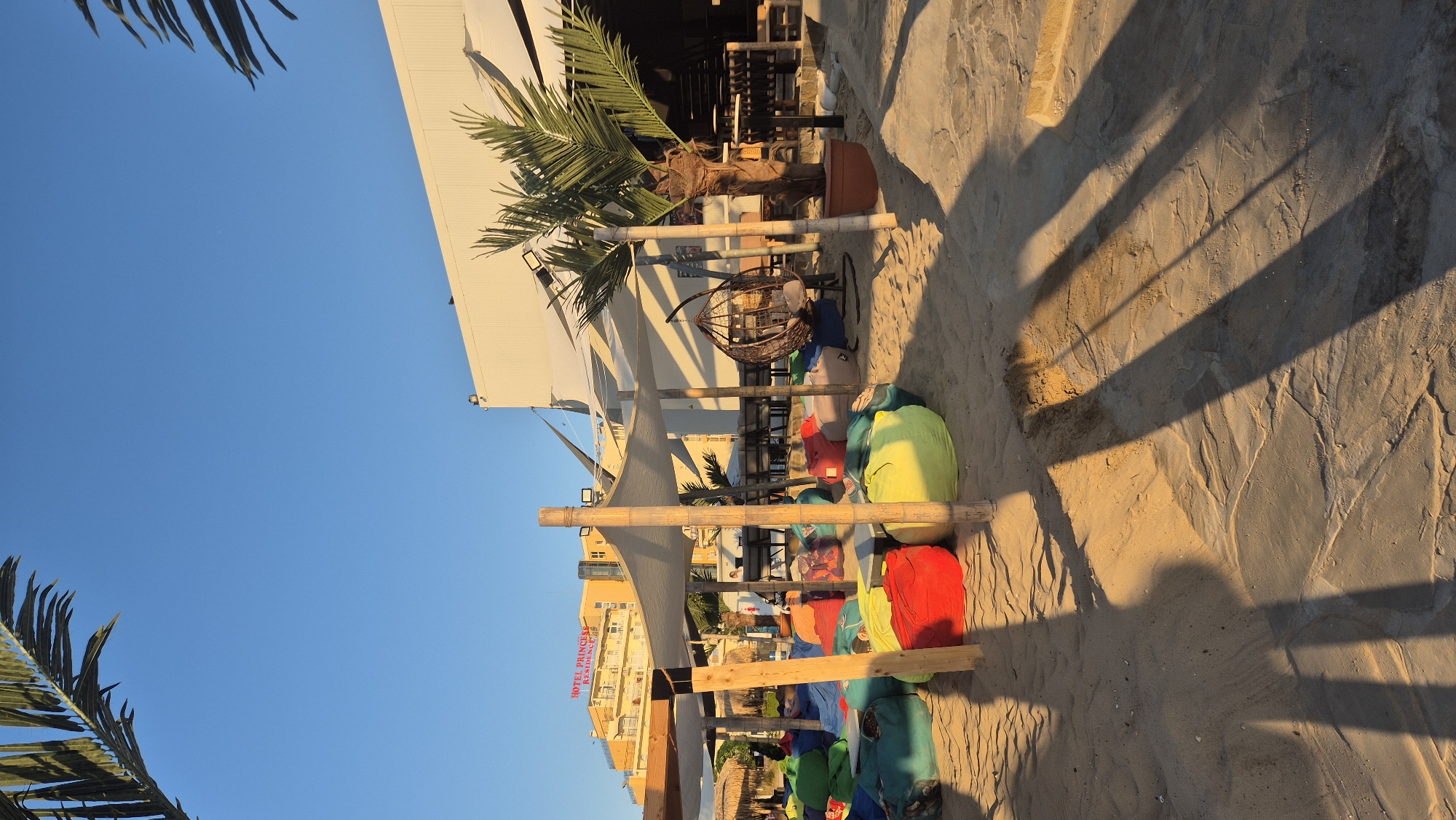 The image depicts a beachside establishment in Kiten, Bulgaria, bathed in the warm glow of late afternoon sunlight. The foreground is dominated by a sandy expanse with numerous colorful beanbag chairs in shades of red, yellow, green, and blue scattered around low tables. These seating arrangements are partially covered by a light-colored, fabric canopy supported by wooden poles. In the middle ground, a suspended woven chair hangs from a stand, inviting relaxation. Behind the seating area, a modern building with white siding and dark windows forms the backdrop. A palm tree in a brown planter stands to the right of the suspended chair, adding to the tropical beach vibe. Further in the distance, a multi-story yellow building with the signage "HOTEL PRINCESS RESIDENCE" is visible. The sky is a clear, bright blue, and shadows from the canopy and palm tree are cast across the sand, indicating the time of day. There are no identifiable people clearly visible, though figures can be discerned in the distance near the hotel and within the establishment. The overall scene suggests a relaxed, open-air dining or lounging area by the sea.