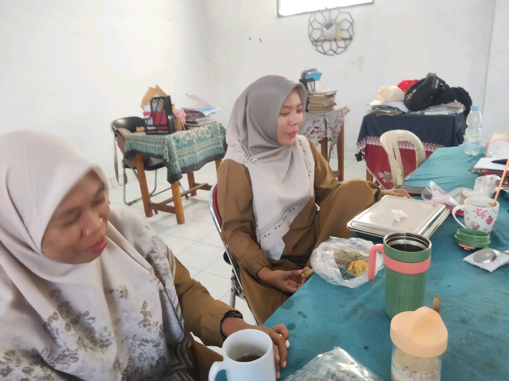 The image depicts two women wearing hijabs and brown uniforms seated at a blue-clothed table in what appears to be an indoor setting. The woman in the foreground is wearing a light-colored hijab with a floral pattern and a brown uniform. Her face is partially visible as she looks down, her lips slightly parted. She is holding a white mug with a dark liquid, and her hand rests on the table. Next to her is a green thermos with a pink band and handle, filled with a dark liquid. Further on the table, there is a plastic bag containing food, a silver serving tray, and a small white cup with a red strawberry pattern.
The second woman, in the background, is wearing a light grey hijab and a similar brown uniform. She is looking to her right, her mouth open as if speaking. Her hands are visible, one resting near a plastic bag of food. Behind the women, there are tables covered with patterned cloths, displaying stacks of books, papers, and other items, including a red object and a black bag. A water bottle is also visible on one of the tables. A white chair with a red trim is positioned between the two women. The walls of the room are white, and a circular decorative item with a mirror is mounted on the wall in the background. The floor appears to be tiled. The overall scene suggests a casual meeting or break in an office or communal space.