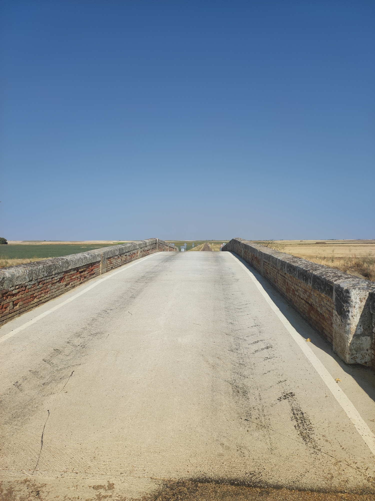 A low-angle, eye-level, full shot captures a concrete road extending into the distance under a clear, bright blue sky. The road is bordered by rustic, weathered brick walls on both sides, topped with stone. The walls show signs of age and some damage. The road surface itself is a light-colored concrete, with some dark tire marks and debris scattered across it, suggesting recent use.

To the left and right of the road, the landscape consists of dry, golden fields, indicative of agricultural land, possibly crops harvested or dormant. The horizon line is low, emphasizing the vastness of the sky. In the far distance, along the road, there appear to be some faint signs or structures. The scene evokes a sense of a rural setting, likely in a warm climate given the dry vegetation and clear sky. The location is identified as Autillo de Campos, Spain. There are no people or animals visible in the image.