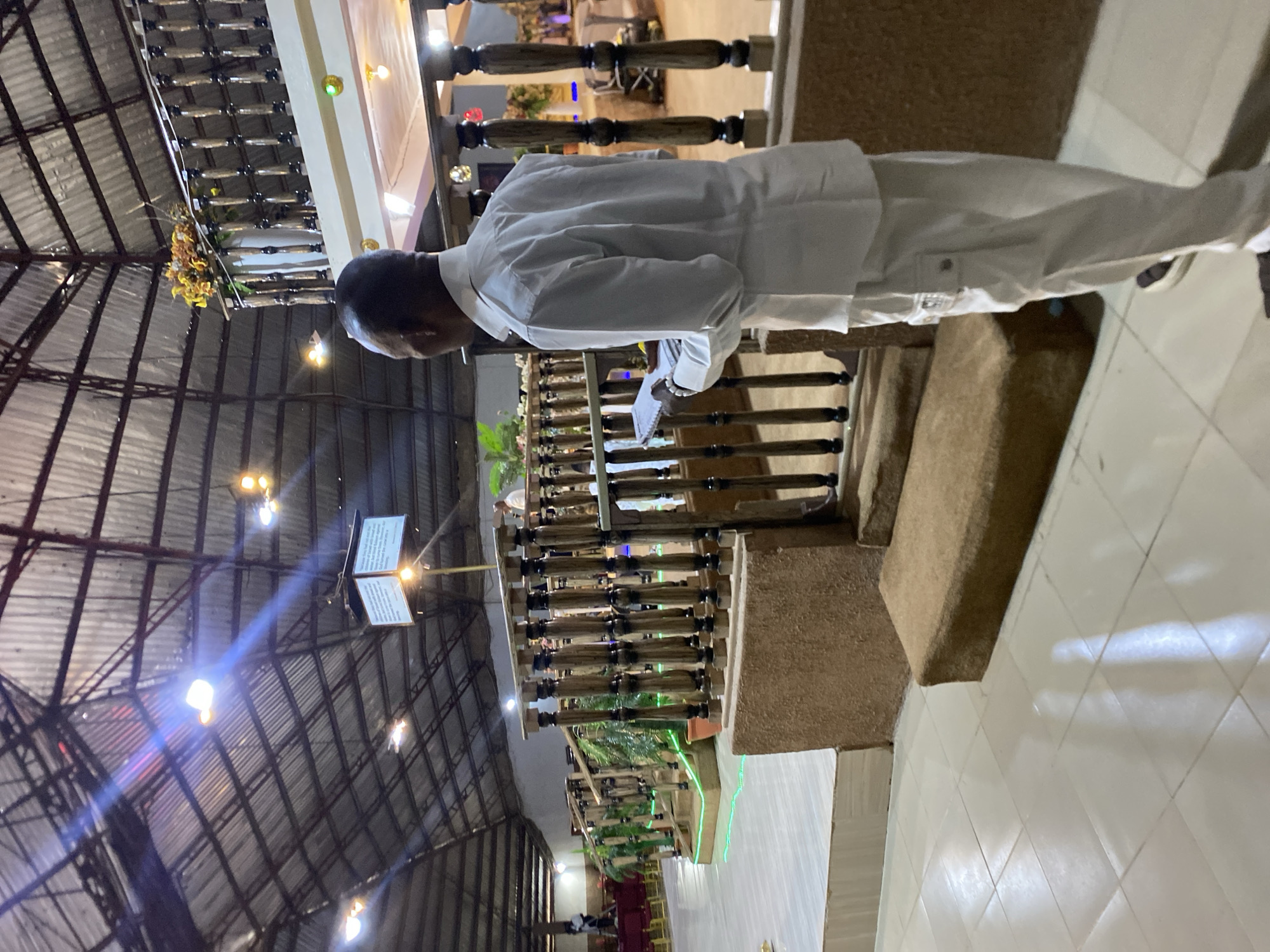 An indoor shot in Jos, Nigeria shows a man wearing a white shirt and pants standing behind a wooden railing reading from a document. The building's high ceiling is supported by metal beams. A display with text is mounted high in the background. The floor is made of white tiles. Green lights illuminate vegetation along the railing below. Chairs are visible in the background.