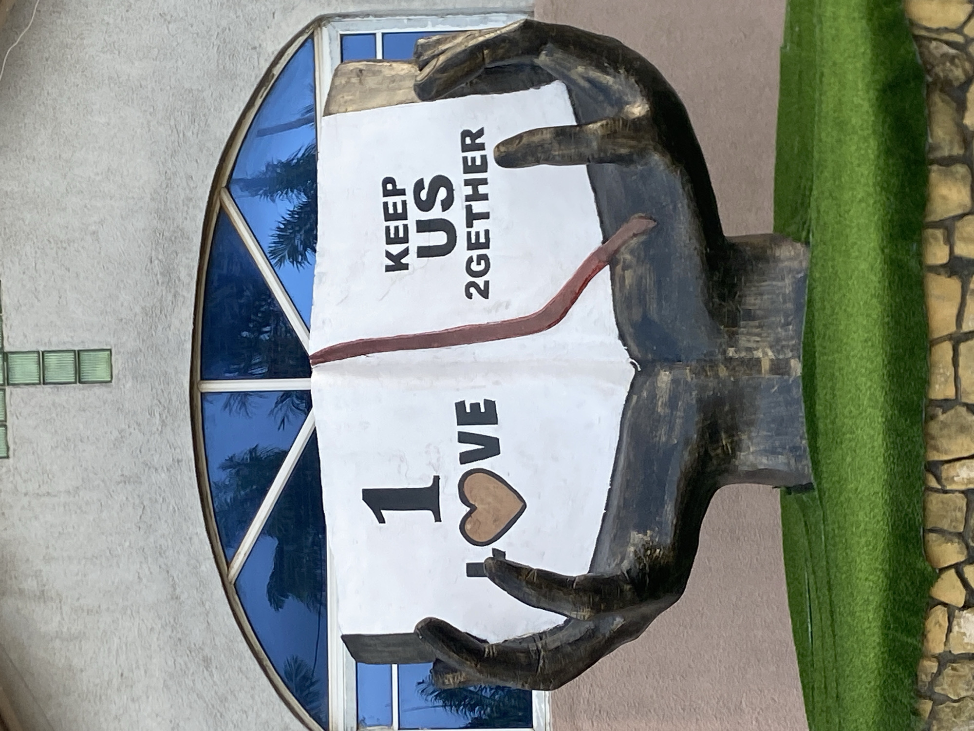 On Rukuba Road, Nigeria, there is an outdoor sculpture of two dark-painted hands holding open a white book. The book has the phrase "KEEP US 2GETHER" on one page, and "I ❤️ VE" on the other. The sculpture is positioned next to a building with a round, multi-pane window with blue-tinted glass. To the right of the sculpture is green grass and a stone-like column. The background building is light gray.