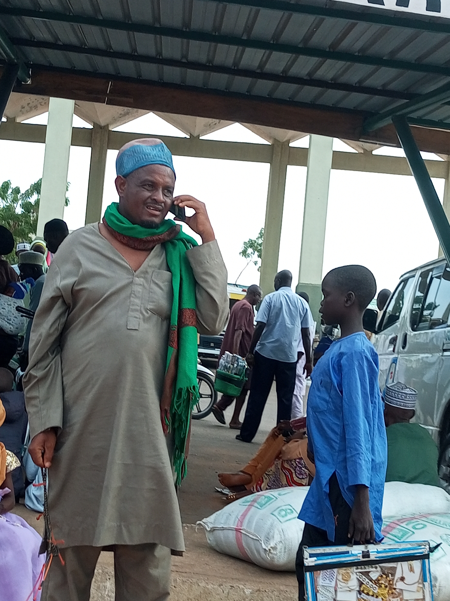 The image depicts a busy street scene in Gombe, Nigeria, under a covered structure with corrugated metal roofing. A man wearing a light brown tunic, a blue cap, and a green scarf is speaking on a mobile phone. He is holding a string of prayer beads in his left hand. To his right, a young boy in a blue tunic stands looking away from the camera. In the background, several other people are visible, some sitting and others walking, along with a silver van. Large sacks are piled on the ground. The overall atmosphere suggests a market or public gathering area.