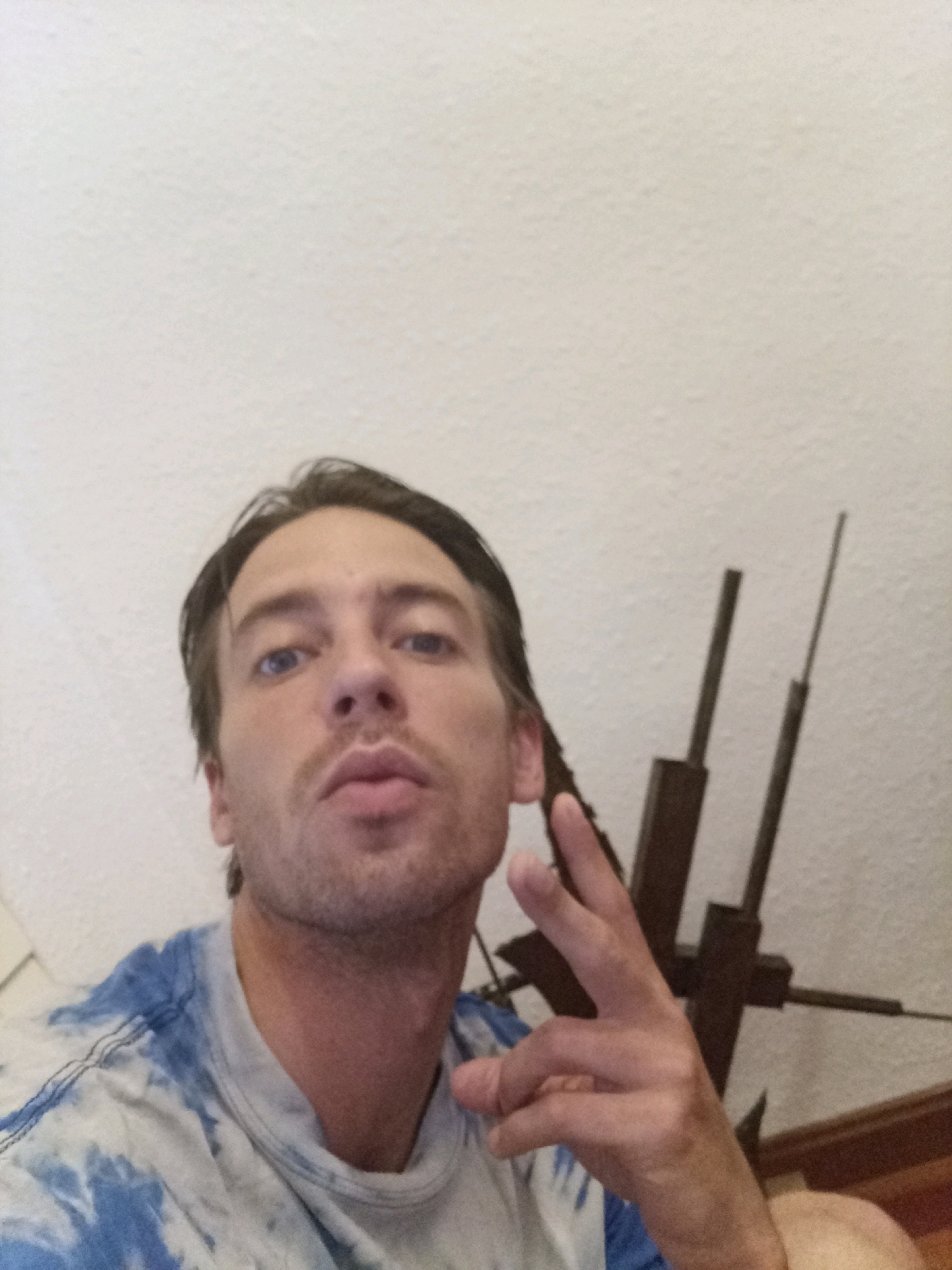 A medium shot, low-angle selfie shows a Caucasian man with brown hair and blue eyes in a tie-dye t-shirt. He is looking at the camera and making a peace sign with his right hand. The background is a textured white wall and what appears to be a dark metal sculpture. The man's left arm and part of his shoulder are also visible. The image appears to have been taken indoors. There is no discernible information in the image to suggest a location in Tarragona, Spain.