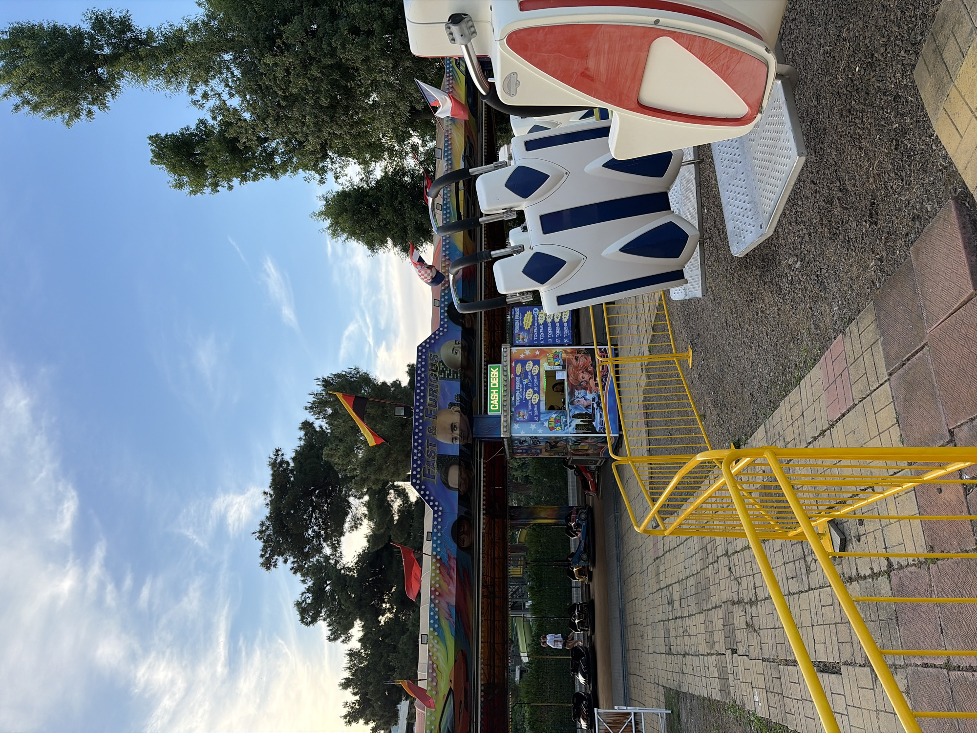 An outdoor amusement park scene is captured with a clear blue sky and scattered clouds. In the foreground, a portion of a white and red amusement park ride is visible, featuring blue and white seating units. Yellow metal railings guide a path through the park. The ground is covered in a mix of gravel and patterned paving stones. Behind the ride, a bumper car attraction is set up, with several bumper cars on the track and a "CASH DESK" booth displaying pricing for tokens. The attraction's overhead structure is decorated with colorful graphics and flags, including images that appear to be from the movie "Fast & Furious." Mature trees line the background, suggesting a temperate climate. A few people are walking near the bumper car area.