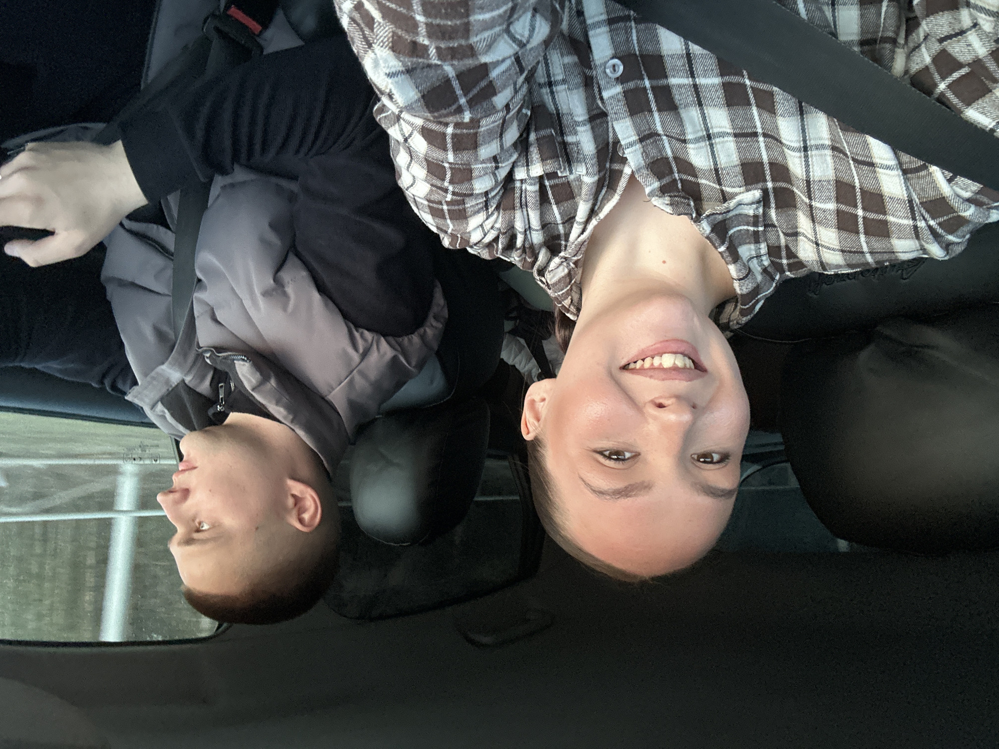 The image is a portrait-oriented photograph taken inside a vehicle, featuring two individuals, displayed inverted (upside down).
If viewed upright, a woman occupies the right side of the frame, captured from the chest up, smiling broadly towards the camera. She has light skin, dark eyes, and her hair appears pulled back. She is wearing a brown and white plaid button-up shirt, and a black seatbelt crosses her chest and shoulder. Her expression conveys cheerfulness and engagement.
On the left side of the frame, a man is seen in profile, looking towards the left out of a car window. He has very short, dark hair and light skin. He is dressed in a dark long-sleeved shirt under a grey padded jacket, and a dark seatbelt is also visible across his chest. His right hand, with light skin, is partially visible in the lower left corner.
The setting is the interior of a car, characterized by dark upholstery and visible headrests behind both individuals. Through the window next to the man, a blurred outdoor scene can be discerned, suggesting movement or a diffused light, possibly indicating overcast weather. There is no visible text in the image. The photograph captures a casual, happy moment during a car journey.