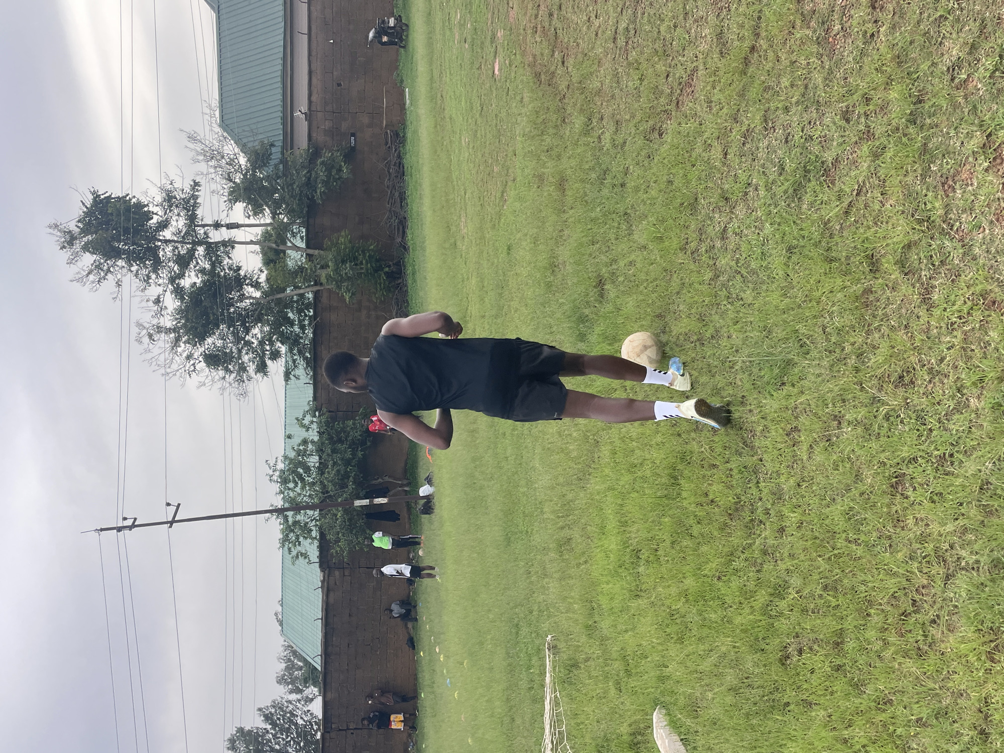 A young Black male, wearing a black t-shirt and shorts, is playing soccer on a grassy field. He is in mid-stride, with a soccer ball at his feet. The field appears to be part of a training ground, with several other individuals scattered around in the background, some of whom also appear to be involved in soccer drills. There are various cones placed on the grass, and a soccer net is visible in the lower right corner.
The setting is outdoors, with a cloudy sky suggesting overcast weather. A brick wall runs along the back of the field, and beyond that, a building with a green roof is visible, along with trees and utility poles with overhead wires. The overall atmosphere seems to be one of athletic activity and training. The time of day is likely daytime, given the lighting conditions. No explicit text is visible in the image. The location context is Jos, Nigeria.