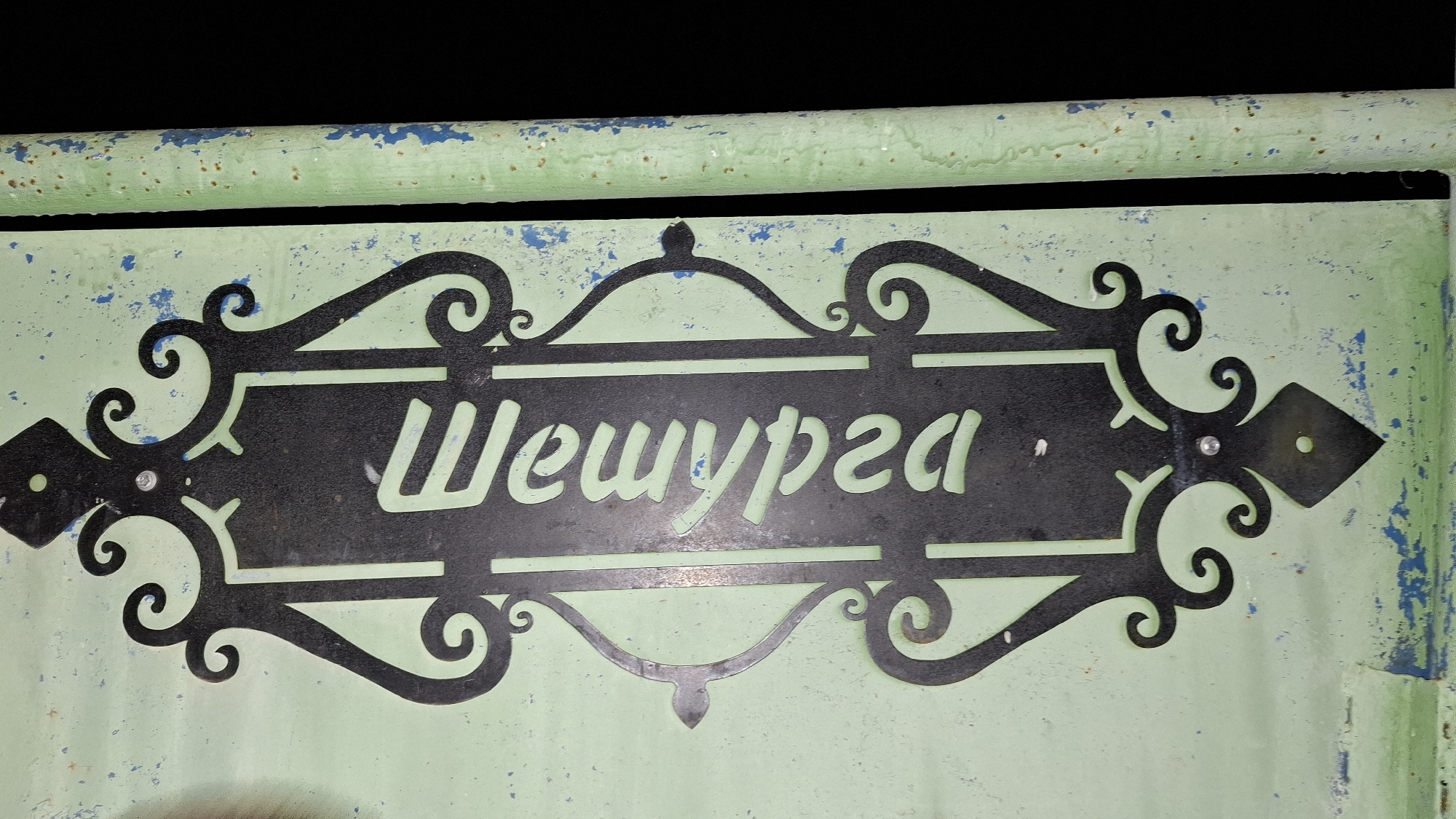 The image shows a sign reading "Шешурга" (Sheshurga) in a decorative black frame mounted on a light green surface with blue paint chipping. The frame has ornate flourishes and diamond-shaped ends, each secured with a visible rivet. The surface appears weathered. The sign is located in the city of Sheshurga, Russia, as indicated by the text on the sign.