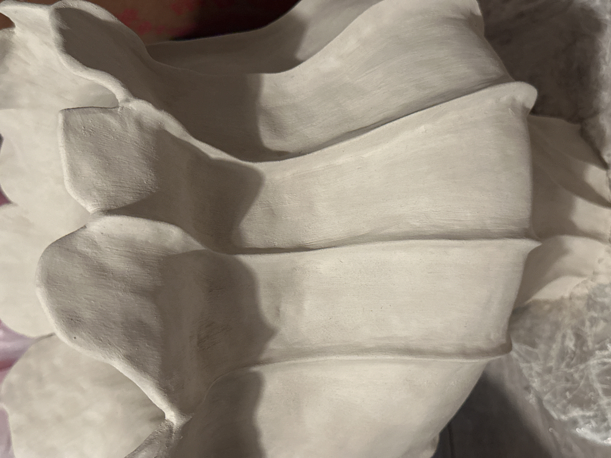 The image shows a close-up of a cream-colored, unglazed ceramic vessel with a scalloped or petal-like exterior. The texture of the ceramic is visible, appearing somewhat rough and porous. The vessel has a distinct fluted design, with vertical indentations creating a series of overlapping sections. Shadows are cast by these indentations, highlighting the three-dimensional form of the object. The background is out of focus, with hints of pink and darker colors suggesting the presence of other objects or a dimly lit environment. There is no identifiable geographical information or context to suggest a location in Nevinnomyssk, Russia. The image focuses solely on the detail of the ceramic object.