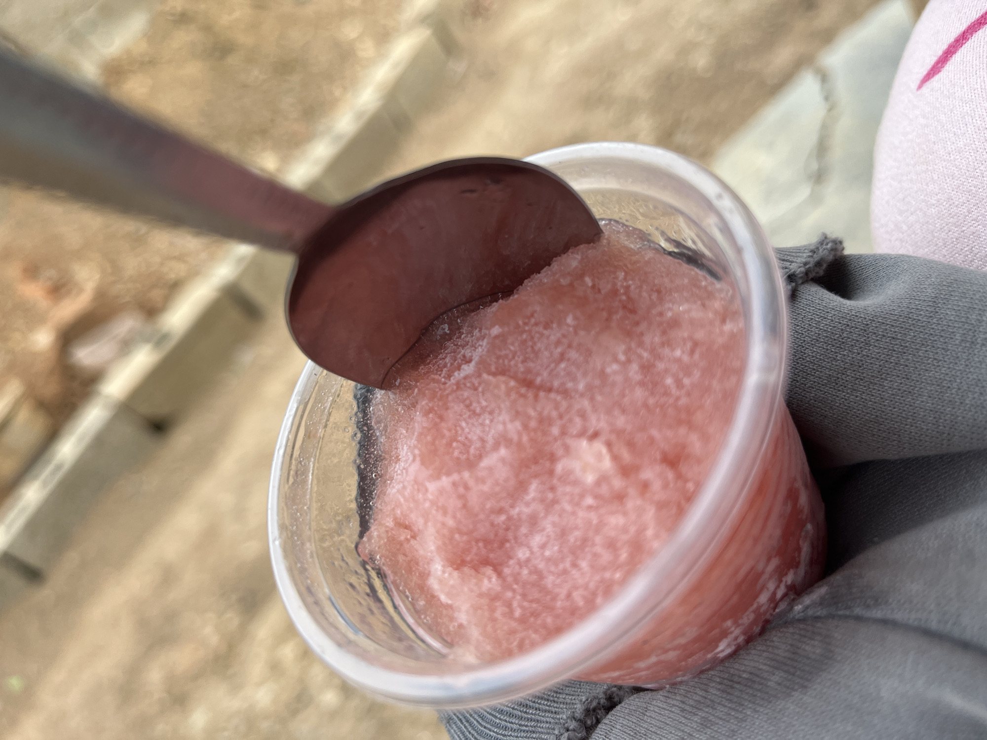A close-up, tilted, high-angle shot features a transparent plastic cup filled with pink slushy granita. A metal spoon is positioned above the cup, partially submerged into the granita. The cup is held by a person whose hand is covered in gray fabric. A portion of a pink and white patterned garment is visible on the right edge of the image. The background suggests an outdoor setting with beige and gray tones. No specific details within the image directly link it to the city of Juan Griego, Venezuela.