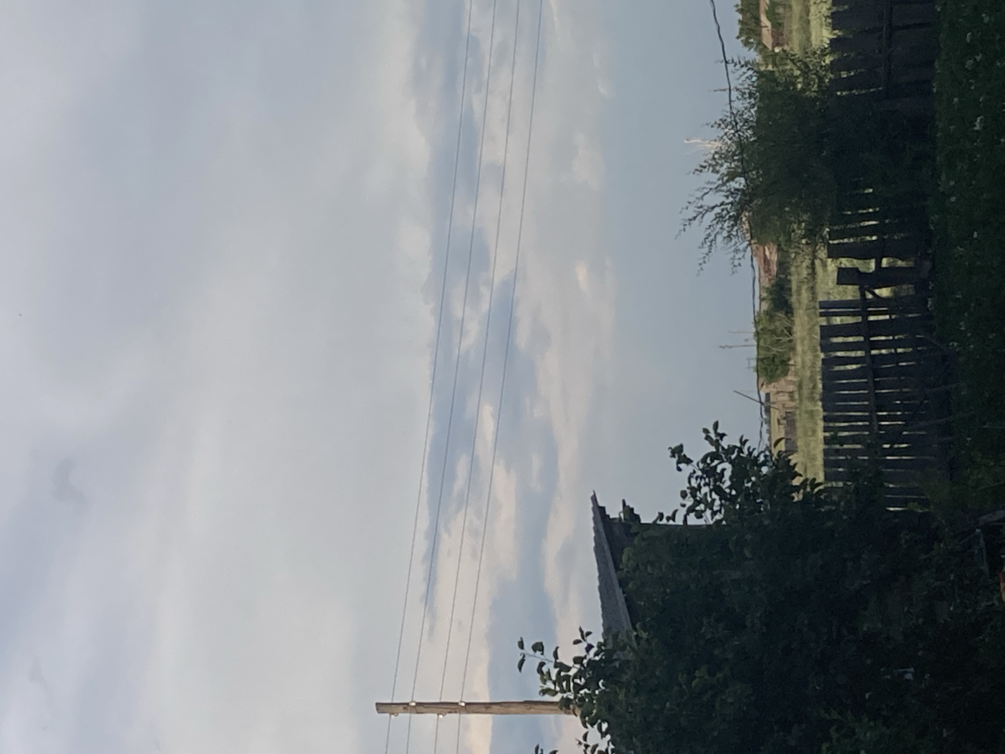 The image depicts an outdoor scene that appears to be taken in a rural setting, possibly in Rakity, Russia. The perspective is angled, making it difficult to discern a clear horizon line. The dominant elements are the sky, a wooden fence, trees, and utility lines.
The sky is largely overcast with patches of lighter and darker clouds. The presence of utility lines, running horizontally across the frame, suggests the proximity of a power source or telecommunication infrastructure. A wooden fence, constructed from horizontal planks, runs along the right side of the image, suggesting a boundary or enclosure. Lush green trees are visible, with one particularly dense tree in the foreground partially obscuring a structure with a dark, angled roof, possibly part of a building or shed.
The lighting suggests it could be daytime, with diffused light due to the cloud cover. There are no people or animals visible in the image. The overall impression is one of a quiet, perhaps slightly neglected, rural environment. The weathered appearance of the fence and the dense foliage contribute to this feeling. There is no visible text in the image.