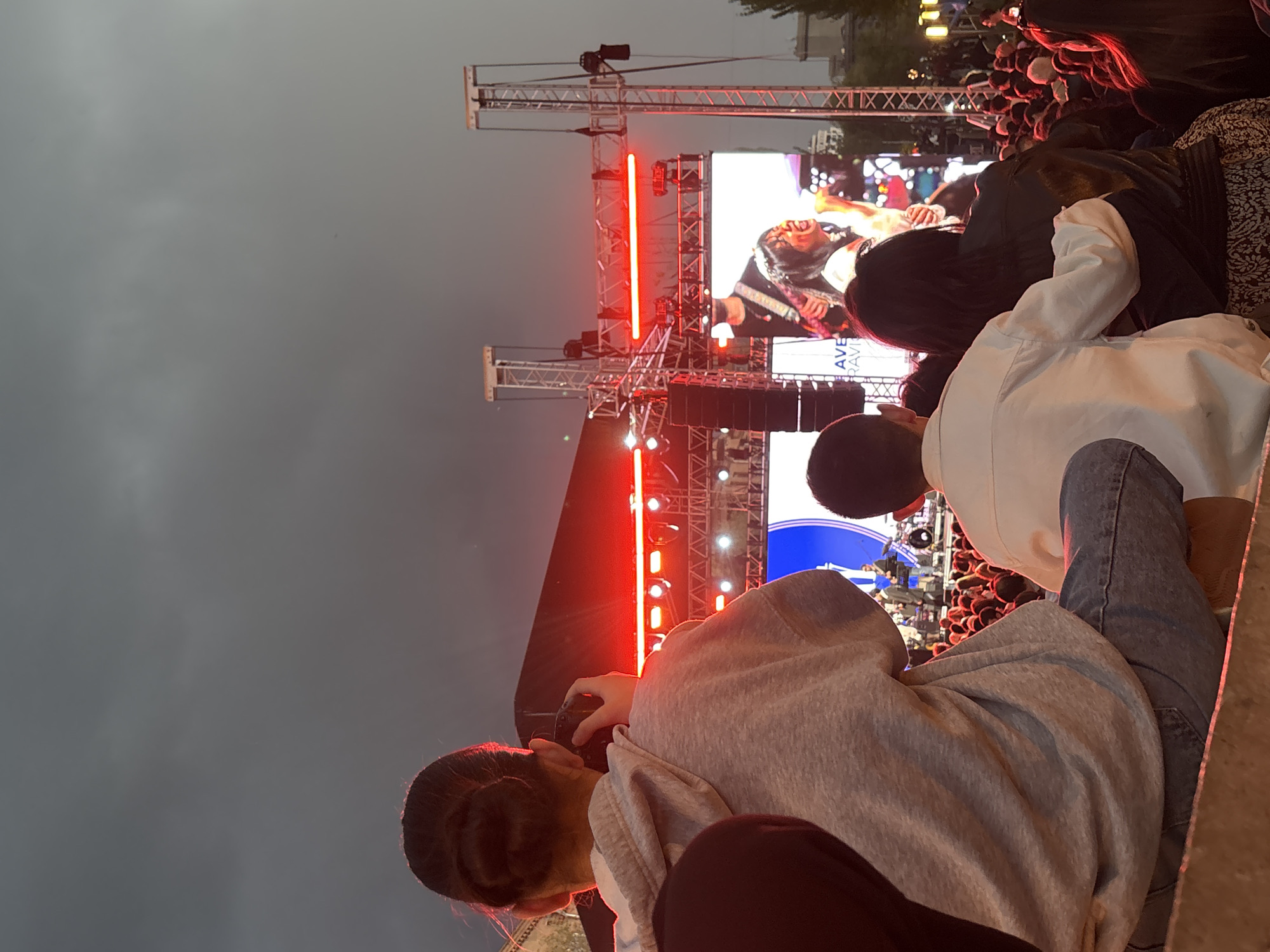 This image captures an outdoor evening event in Yerevan, Armenia, likely a concert or festival. The scene is dominated by a brightly lit stage with large LED screens and metal truss structures against a dark, overcast sky.
In the foreground, two individuals are prominent, viewed from behind. On the left, a person with dark hair pulled back in a bun wears a light grey hoodie and holds a camera, seemingly capturing the event. To their right, another person with short dark hair is dressed in a white shirt and jeans, also facing the stage. Behind them, a crowd of attendees is visible, with many heads and shoulders, suggesting a large gathering.
The stage features multiple large LED screens displaying a performer, who appears to be an energetic singer or musician with long dark hair, an open mouth, and possibly holding a guitar-like instrument, characteristic of a rock or pop performance. The stage is illuminated with intense red lights, casting a glow on the structures, along with other white lights and speakers.
The time of day is clearly dusk or night, indicated by the dark sky and the reliance on artificial stage lighting. The overall atmosphere appears lively and engaging, with the audience focused on the performance. On one of the screens, partial text "AVE" and what looks like "RAVE" is discernible.