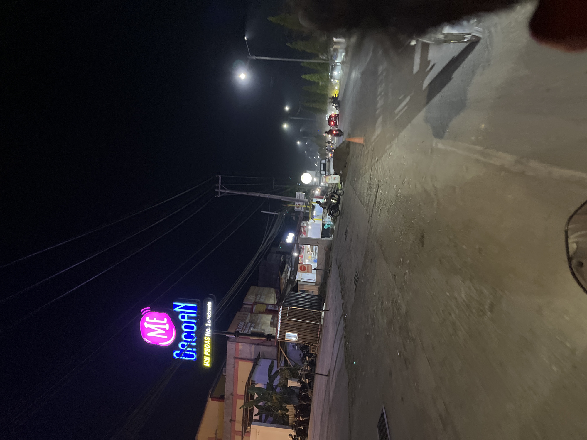 The image depicts a nighttime street scene in Bundar, Indonesia. A prominent neon sign for "ME Gacoan" with the tagline "MIE PEDAS No. 1 di Indonesia" is illuminated in vibrant pink and blue. The street is paved and appears wet, reflecting the lights from streetlamps and nearby buildings. Several motorcycles are parked on the left side of the road, and a few are in motion on the street. A red car is also visible in the distance, along with other vehicles. Traffic cones are placed on the side of the road, suggesting ongoing construction or road work. The environment is characterized by the dark night sky, illuminated by artificial lights, and the presence of typical urban infrastructure like streetlights and power lines.