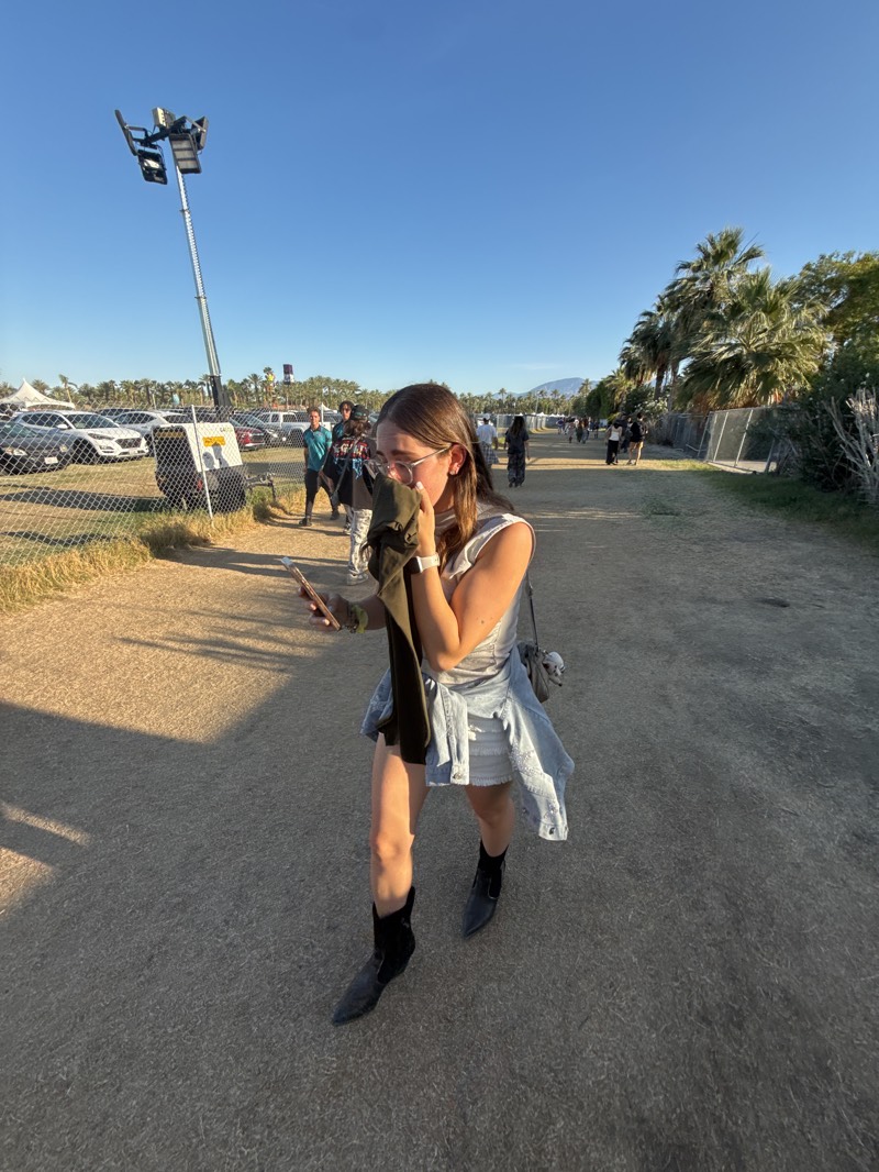 The image captures a person walking on a path with palm trees on the right side, under a clear blue sky, likely in Indio, United States. The individual is wearing a white top, denim skirt, black boots, and a light denim jacket tied around their waist. They are also holding a phone in one hand and a dark-colored item in the other. To the left of the path, there's a fenced-off area with parked cars and a light tower. People are seen walking in the distance along the path. The ground appears to be a dirt or gravel surface.