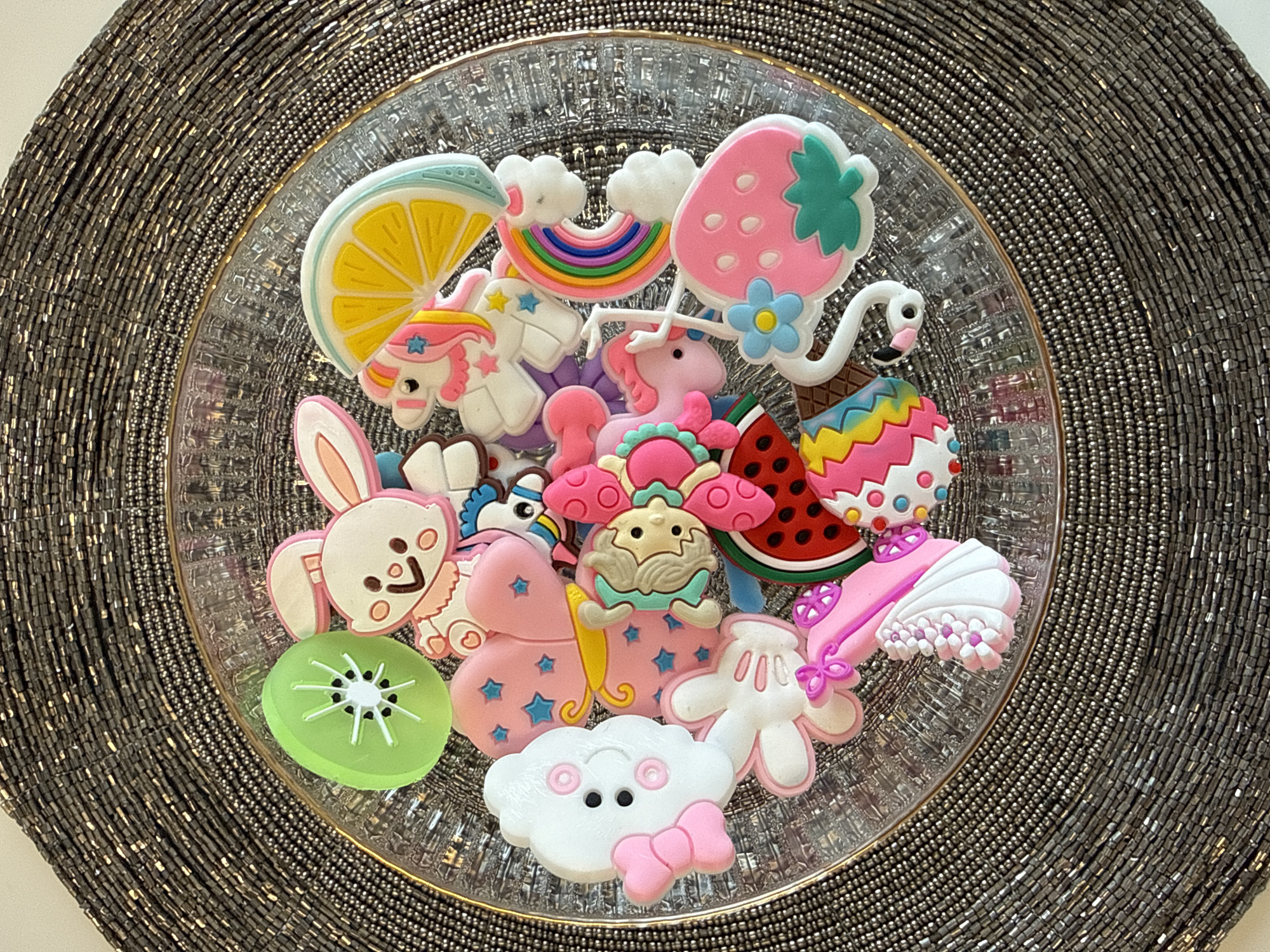 A top-down, close-up shot shows a clear glass plate filled with colorful novelty items, centered on a beaded circular placemat. The plate contains a collection of playful, cartoonish trinkets: a slice of lemon, a rainbow with clouds, a strawberry, a flamingo, an ice cream cone, a slice of watermelon, a bunny, a kiwi slice, two pink unicorns, a creature with a tan face, a pink butterfly with stars, a white cloud with a pink bow, and Mickey Mouse's white gloved hand. The placemat has a metallic silver-gray color. The background consists of a white surface, likely a table. There is no indication of the location being in Votkinsk, Russia.