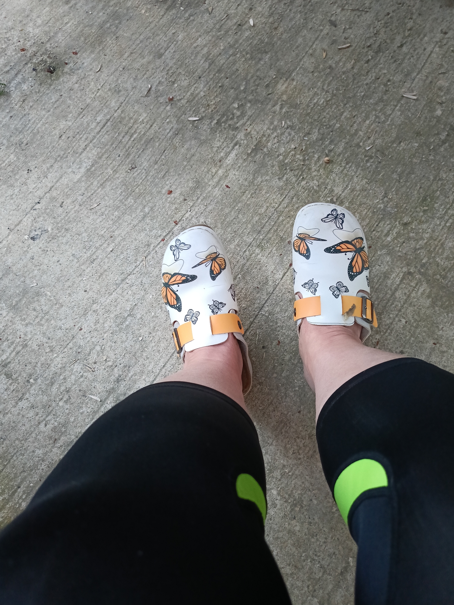 A person's feet wearing white clogs with orange and black butterfly designs are visible, standing on a concrete surface. The person is wearing black pants with bright green accents on the sides. The concrete appears to be outdoors, possibly in a walkway or patio area, with scattered debris. The image does not provide any information about a specific location in Varna, Bulgaria.