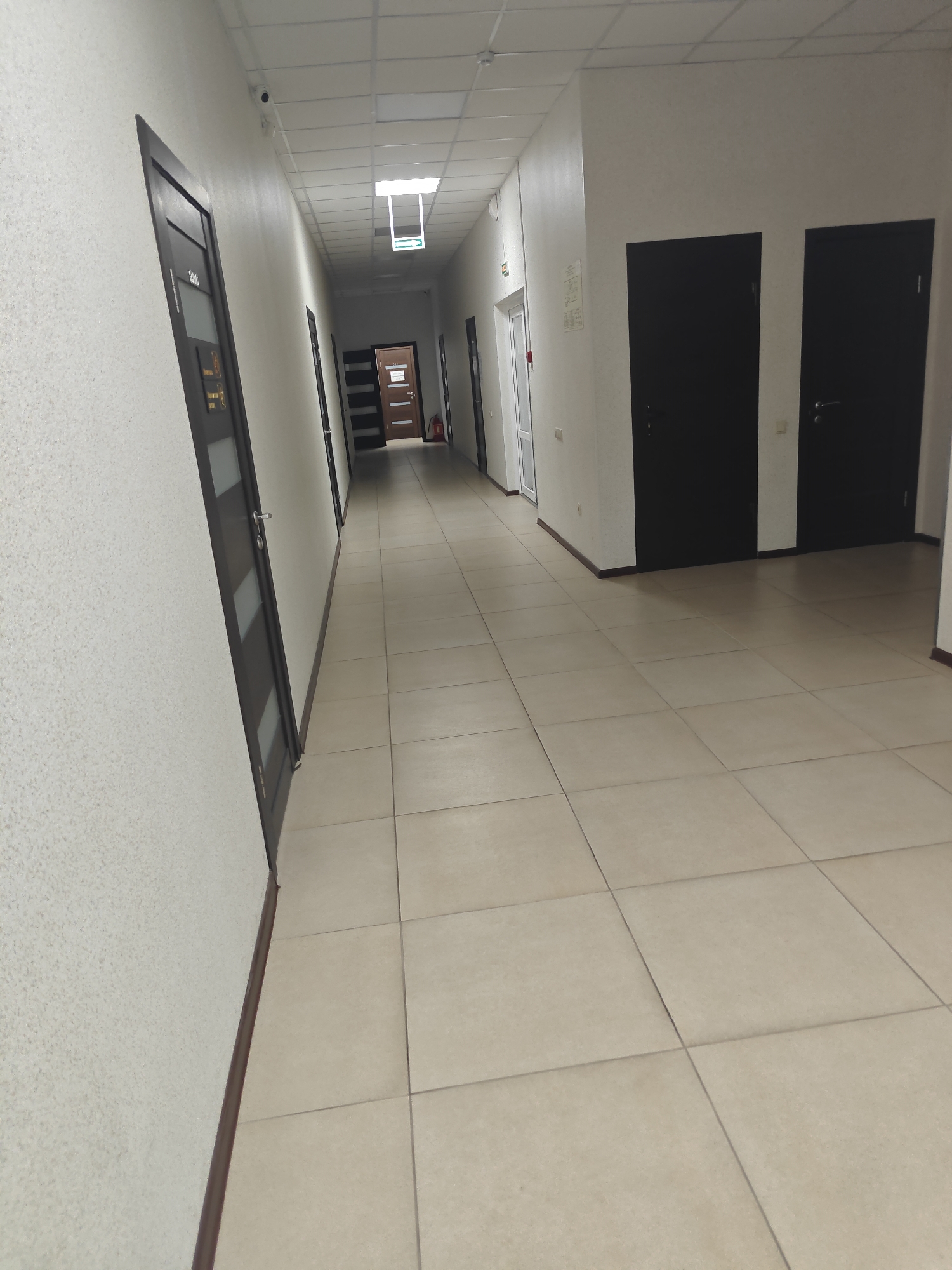 A long, tiled hallway with multiple doors. The floor is covered in light beige square tiles. On the left wall, there is a textured white finish, and several dark doors with glass inserts are visible. On the right wall, there are two large, dark solid doors. At the end of the hallway, more doors are visible, including one with a wooden finish. A ceiling light fixture with a green exit sign is visible. A fire extinguisher is also present. The scene appears to be an interior hallway, likely in an office building or similar institutional setting. No people are visible in the image. The location is not specified in the image.