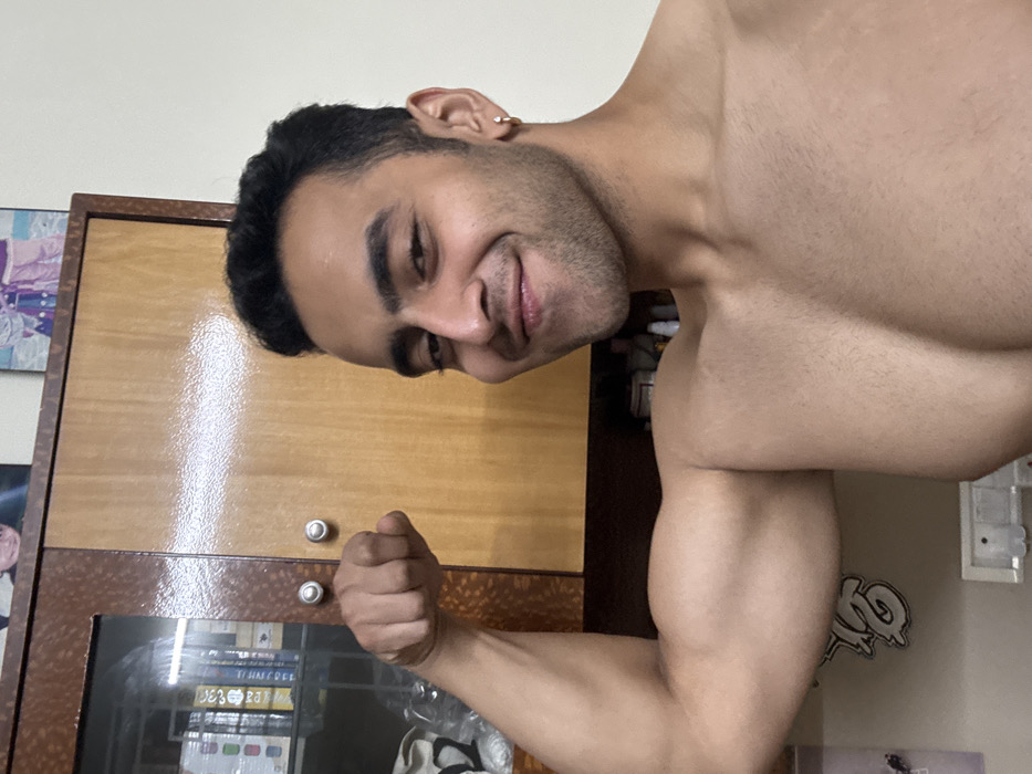 A shirtless man with dark hair and a small earring flexes his bicep, gazing downwards with a subtle smile. He stands close to a wooden cabinet with glass doors. A framed image hangs on the wall. The overall composition has a casual, self-assured vibe, potentially a selfie taken indoors. The image is sideways.