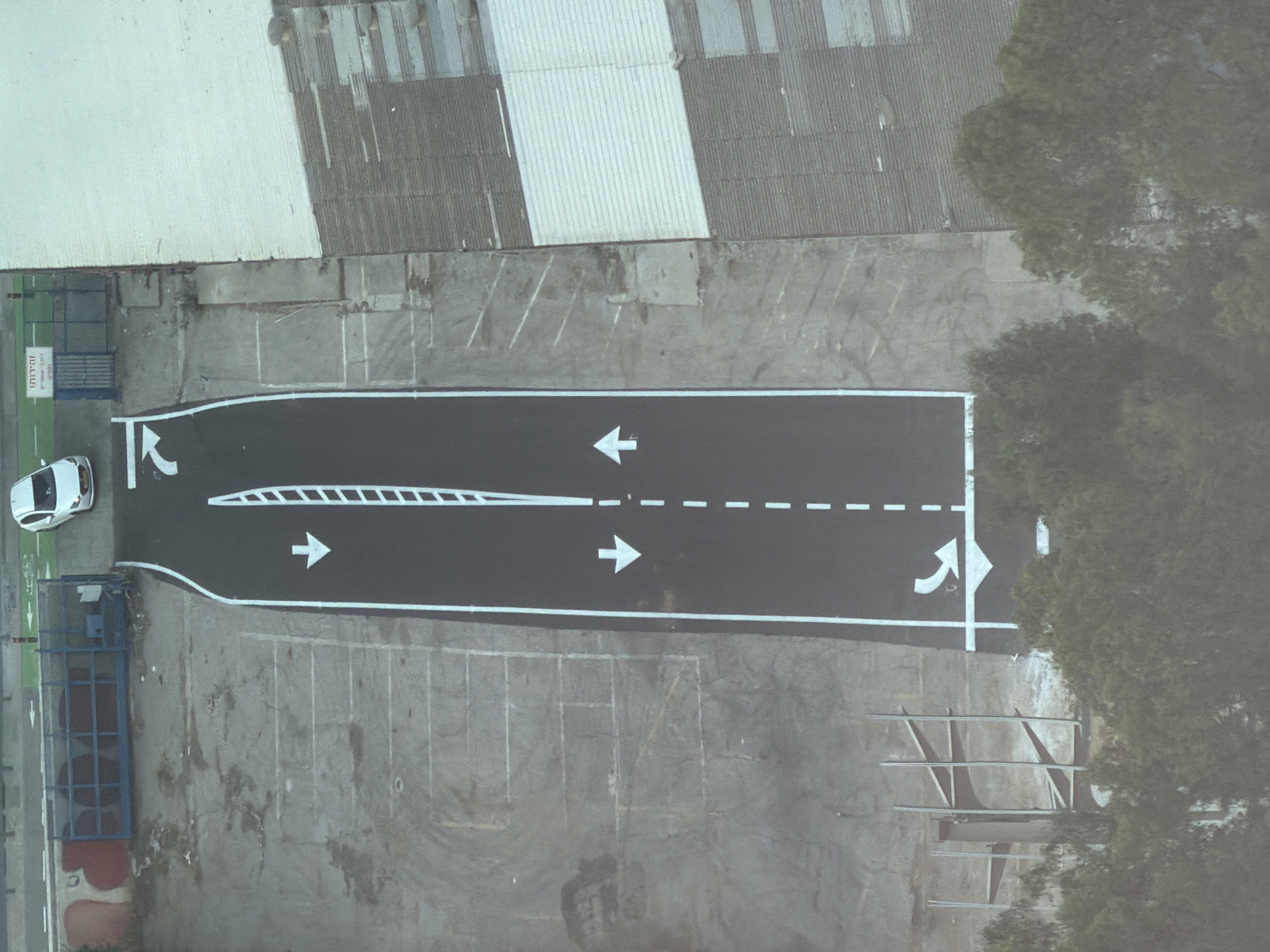 This aerial image depicts a narrow, black asphalt road with white markings, likely a test track or maneuvering area, set within an urban industrial landscape. The road is bordered by grey concrete surfaces and what appears to be a large, light-colored warehouse or industrial building on the right. On the left, there's a grey concrete area with faded white parking lines and a blue metal fence with some graffiti on it. A white car is visible at the top of the image near the entrance to the fenced area, suggesting it has just entered or exited the site. The road itself features white arrows indicating direction of travel, with a single lane in each direction separated by dashed white lines. A vertical element with horizontal white bars runs down the center of the road, possibly a divider or a measurement marker. The background on the left is a rough, grey rock face or embankment, with some foliage obscuring the lower portion. The scene appears to be devoid of people. The presence of a white car at what looks like an entrance gate and the road markings suggest a controlled environment for vehicle testing or training.
While the image itself does not contain explicit markers of its location, based on the visual cues of industrial buildings, urban setting, and specific architectural styles often found in the region, and common knowledge of Tel Aviv's urban development patterns, this scene is consistent with areas within Tel Aviv, Israel that are zoned for industrial or logistics purposes, such as near the Tel Aviv Port area or industrial zones within the greater metropolitan region.