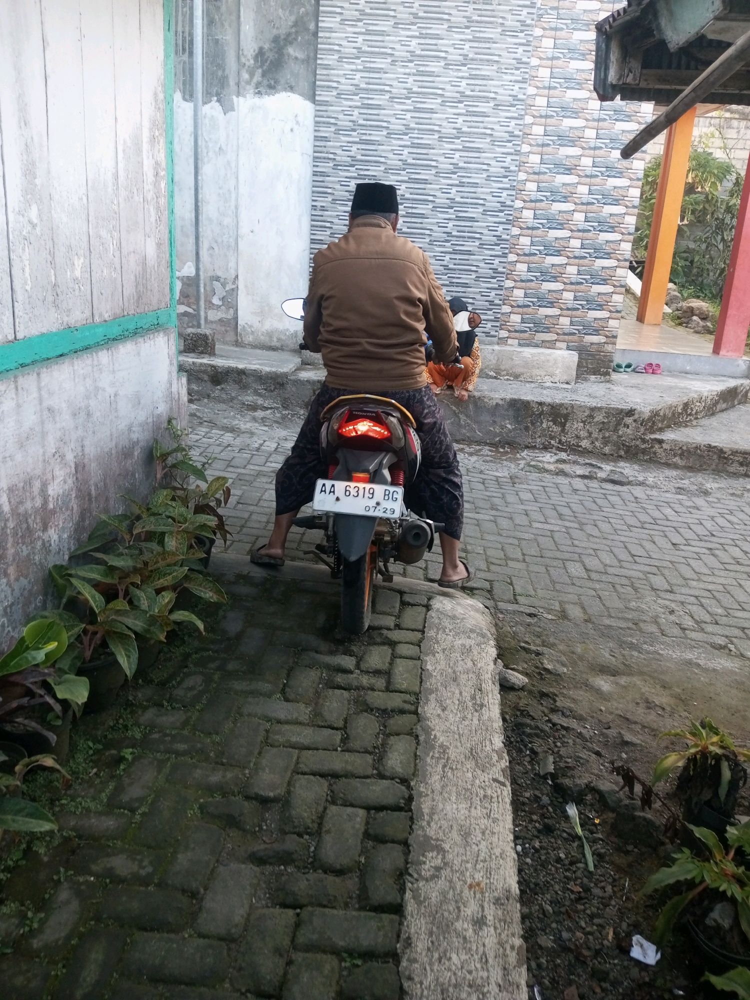 A person wearing a brown jacket and black and white patterned pants sits on a motorcycle. The motorcycle has a license plate that reads "AA 6319 BG". The motorcycle's taillight is illuminated. In front of the motorcycle, a woman wearing a brown and orange patterned dress sits on a concrete step. To the left of the motorcycle, there are several potted plants. The ground is paved with interlocking bricks. The scene appears to be in a residential area.