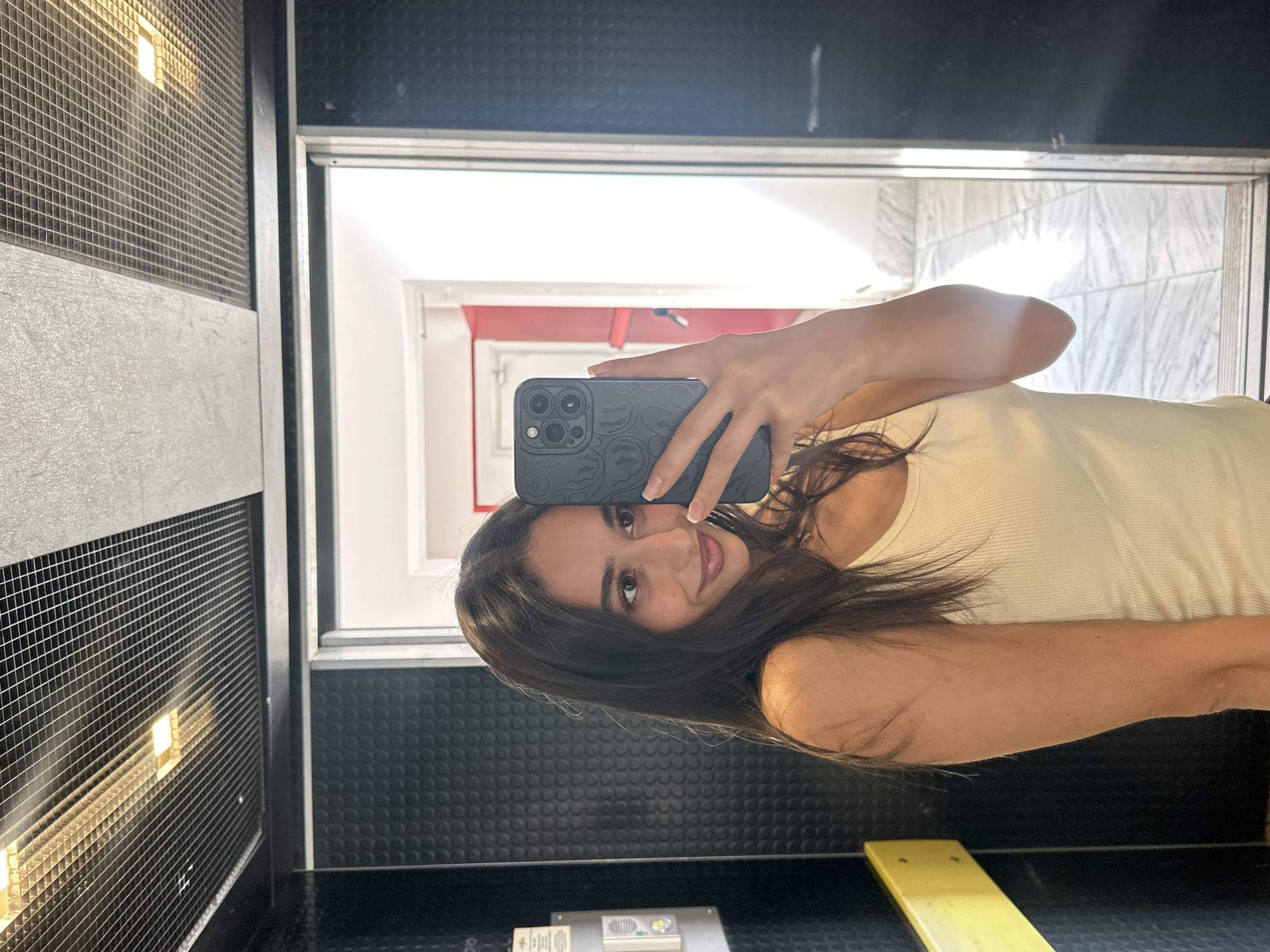 A young woman with long, dark hair is taking a selfie in an elevator. She is wearing a light yellow ribbed tank top and is holding a dark-colored phone with a patterned case in her right hand. Her left arm is slightly bent, and her hand is visible near her face. The elevator interior features black textured walls and a metal ceiling with integrated lighting. A yellow handrail is visible on the left wall. The elevator doors are open, revealing a hallway with red and white walls and marble-tiled flooring. The image appears to be taken in Milan, Italy.