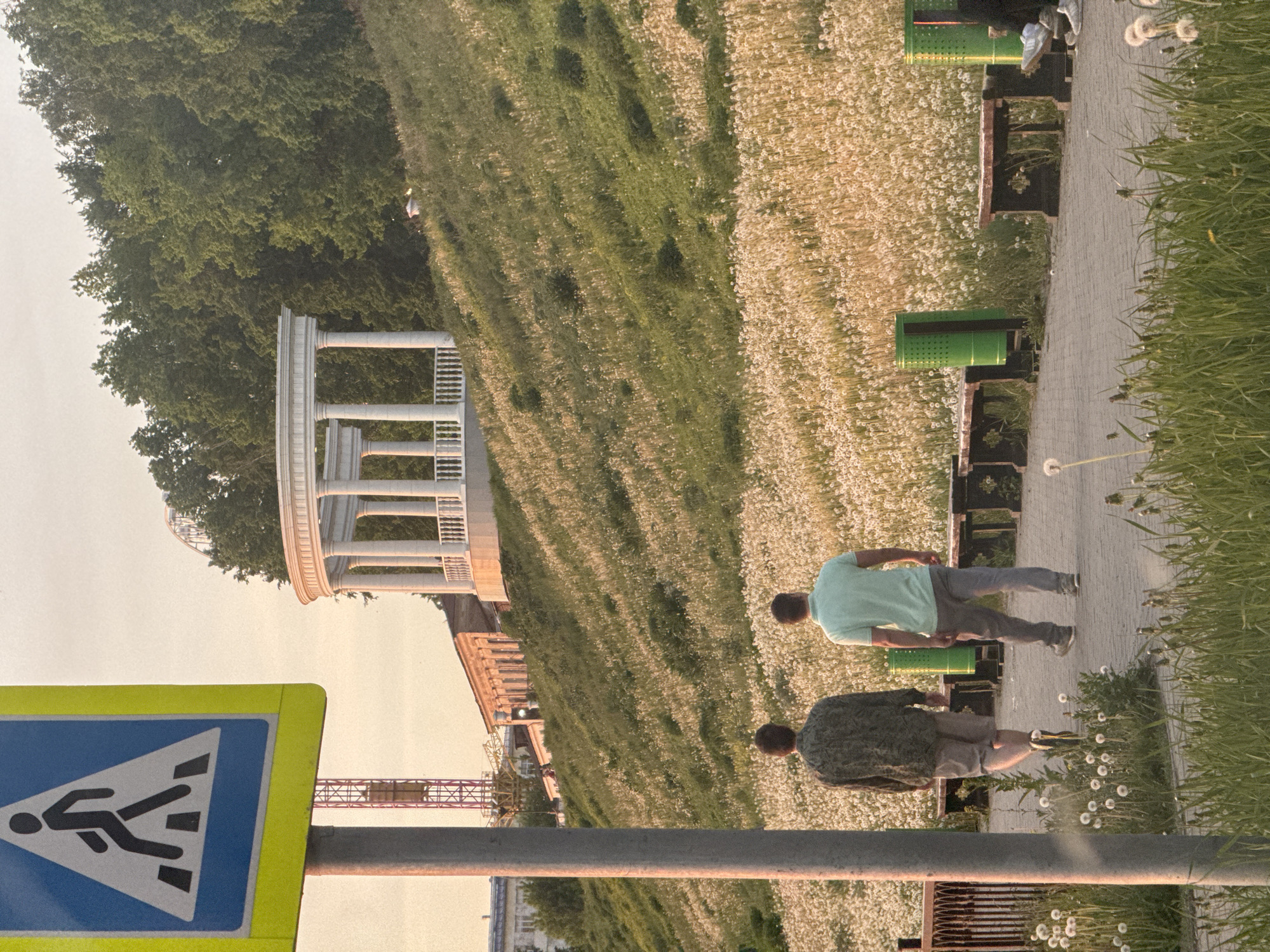 The image captures a scene in Izhevsk, Russia. Two people walk along a paved path bordered by a field of dandelions. The path is lined with benches and flower pots. A white rotunda structure is visible in the background, surrounded by greenery. To the left, a pedestrian crossing sign is prominently displayed. A construction crane is seen in the distance. The overall environment suggests a park or public space.
