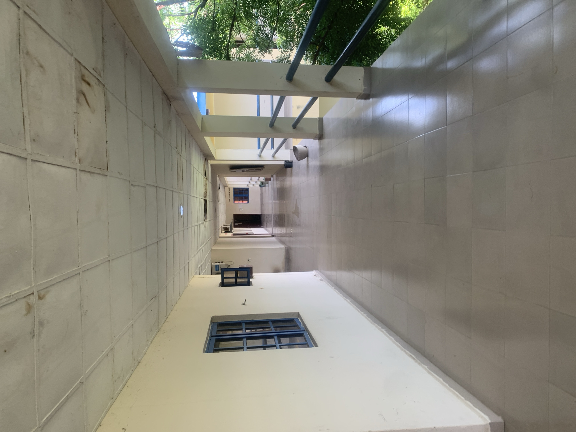 The image depicts an outdoor hallway or covered walkway of a building, likely a school or office complex. The walkway has a tiled floor and a drop ceiling made of square panels, some of which appear to be stained or discolored. On the left side, there is a cream-colored wall with a blue-framed window. Further down the hallway, there are multiple doors and more windows. The right side of the walkway is open to the exterior, featuring support columns and a metal railing overlooking lush green trees. The lighting suggests it is daytime. The image doesn't contain any people or specific actions. The environment appears to be functional but showing signs of age and wear. It is plausible this location is in Jalingo, Nigeria, given the architectural style and surrounding vegetation.