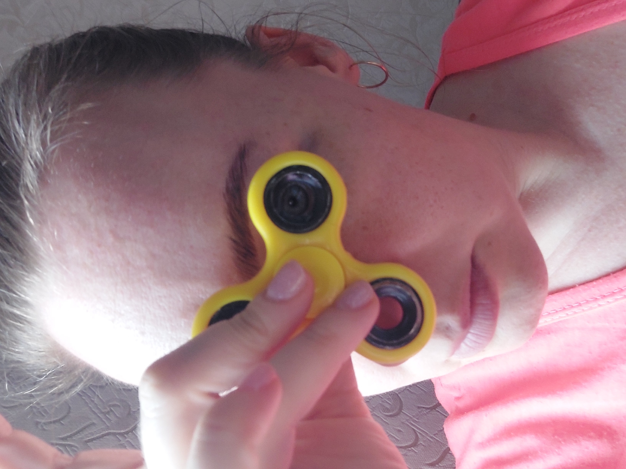 A close-up, waist-up shot shows a fair-skinned person with dark eyebrows and shoulder-length auburn hair, seen from the chest up, with their face turned slightly to the right. They are wearing a bright pink t-shirt and have a small gold hoop earring in their right ear. Their left hand is raised, holding a yellow fidget spinner, positioned in front of their left eye, obscuring it. The background consists of a textured, beige wall on the right and a darker, patterned wallpaper on the left. The lighting is bright, creating a slight glare on the forehead. The image does not provide specific location details within Большой Букор, Russia.