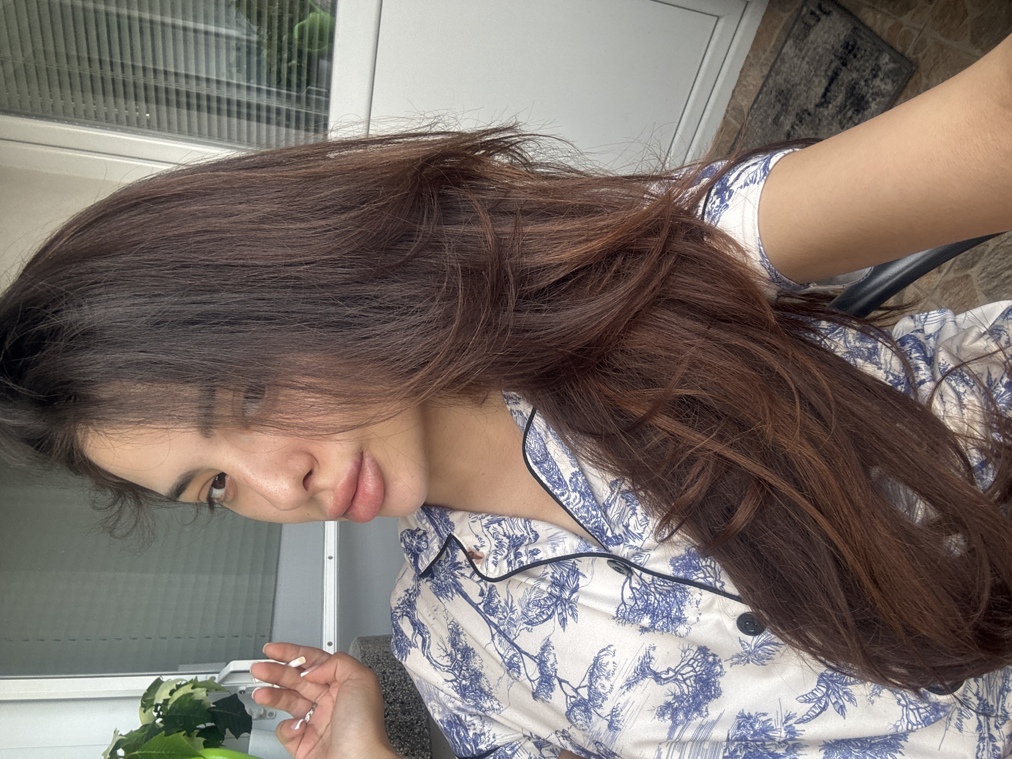 A young woman is captured in a close-up, selfie-style shot, seated indoors near a window. She has long, dark brown hair that cascades over her shoulder and down her chest. She is wearing a blue and white patterned pajama top. Her left hand is raised near her face, and she appears to be holding a cigarette. The background shows a window with blinds, suggesting an indoor setting, possibly a home or hotel room. There are no discernible landmarks or context that would place this image specifically in Plovdiv, Bulgaria.