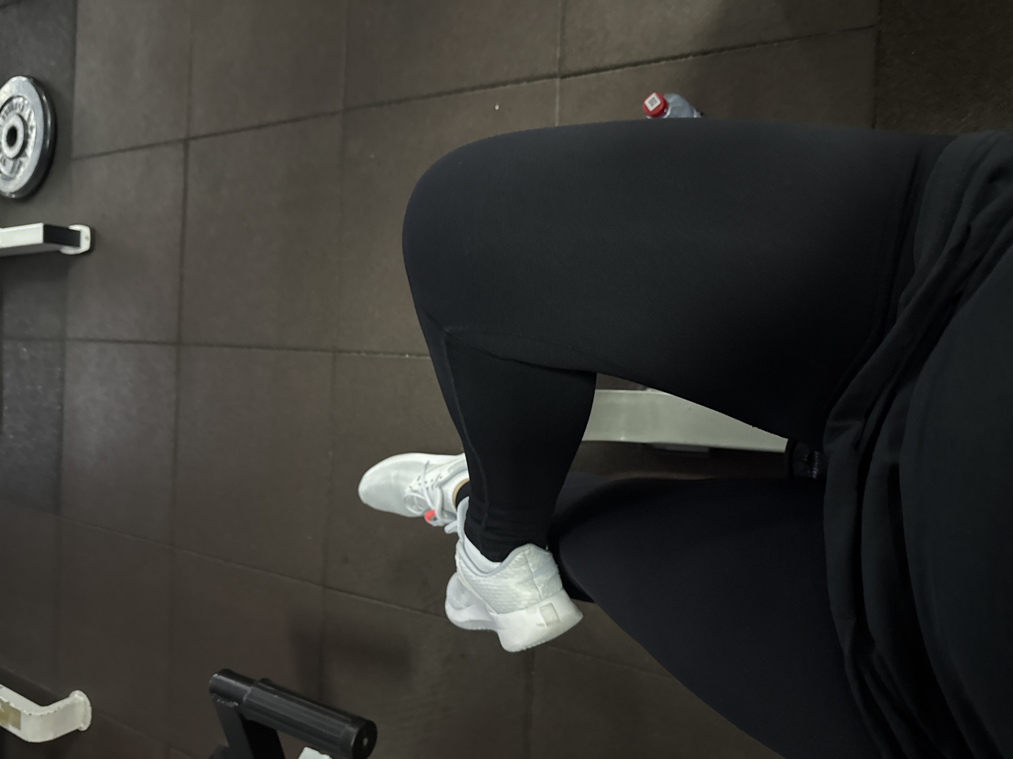The image shows a close-up view of a person's legs and feet in a gym setting. The person is wearing black leggings and white athletic shoes. Their legs are positioned on a piece of gym equipment, possibly a leg press machine, though the equipment itself is mostly out of frame. The background consists of dark gray tiled flooring, common in fitness facilities. In the upper left corner, a weight plate is visible, resting on a bar or rack, further indicating a gym environment. A water bottle with a red cap is also present in the upper right quadrant of the image. The lighting appears to be artificial, typical of indoor gym lighting. There is no visible text in the image. The scene is static, with no clear indication of a specific time of day or weather conditions. The focus is on the lower body of the individual and the immediate surroundings within the gym.
