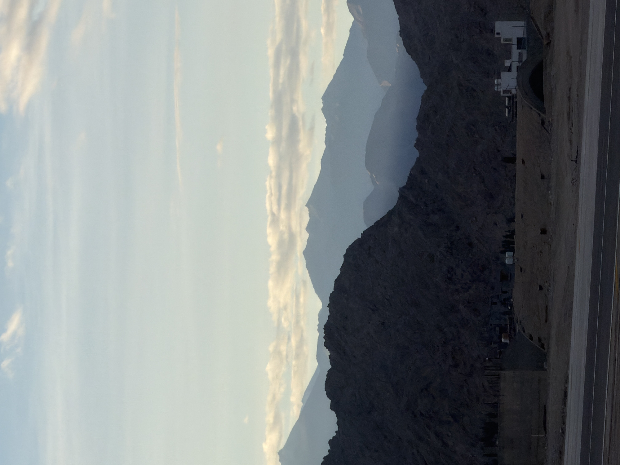 This photograph, taken in the city of Leh, India, depicts a series of silhouetted mountain ranges receding into the distance under a pale blue sky with scattered clouds. The foreground features a dark, rocky mountain slope. Below this, a construction site is visible, with concrete structures, scaffolding, and some equipment. A modern white building with multiple windows and a domed entrance is situated to the right of the construction area. A paved road or highway runs across the bottom of the image, with distinct yellow lines marking the lanes. The scene is characterized by soft, diffused lighting, suggesting it was taken during either dawn or dusk. There are no people visible in the image.