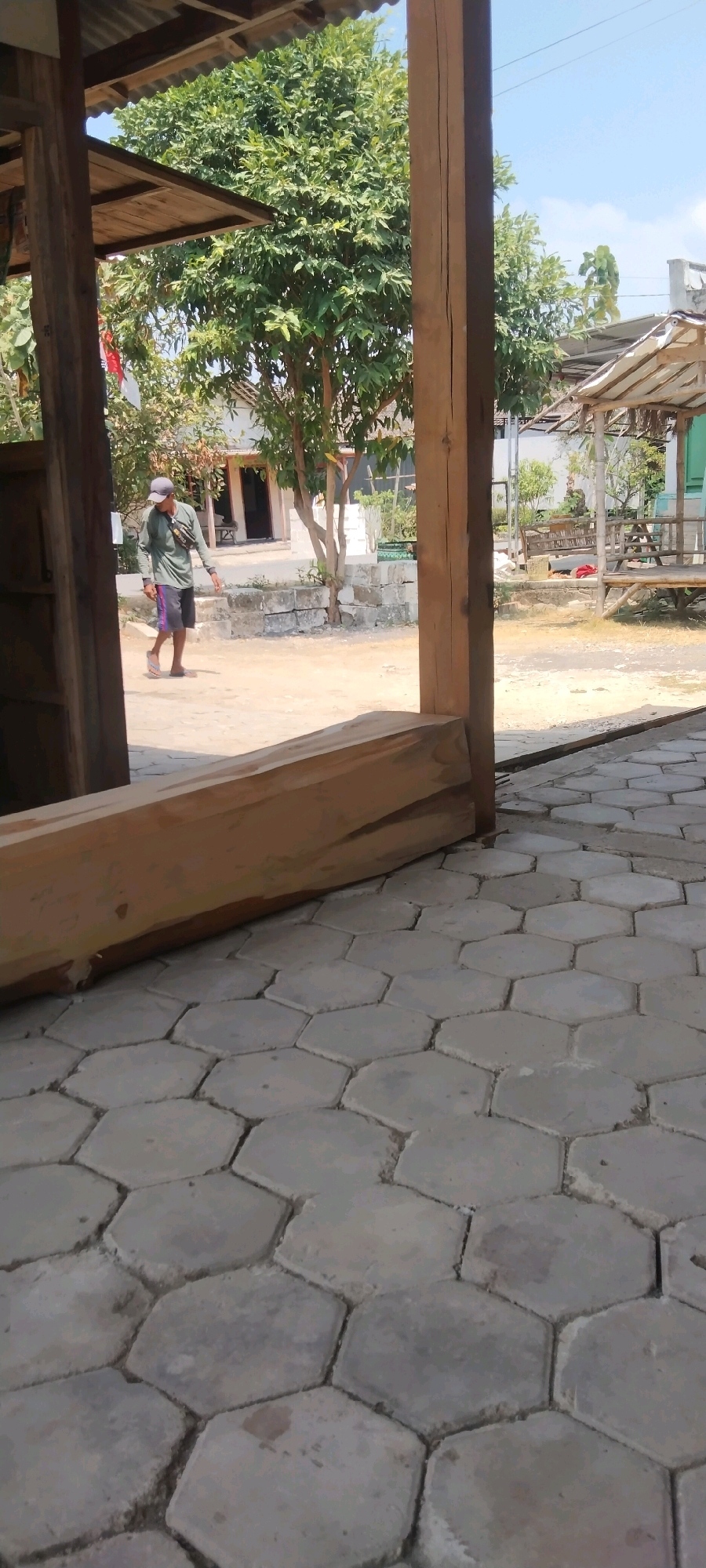 In an outdoor scene, a man walks across a dirt path in what appears to be a village in Indonesia. He is wearing a green long-sleeved shirt, dark shorts, a grey cap, and flip-flops. A black bag is slung across his chest. The man is walking away from the camera, towards a building with an open doorway. To the left of the man, a red and white flag is attached to a pole. Behind the man, a building with a white facade and a brown roof can be seen, surrounded by trees. To the right of the man, another building with a thatched roof and a wooden bench is visible. The foreground of the image is occupied by hexagonal paving stones. The sky is clear and blue, indicating a sunny day. The overall impression is of a rural, tranquil setting.