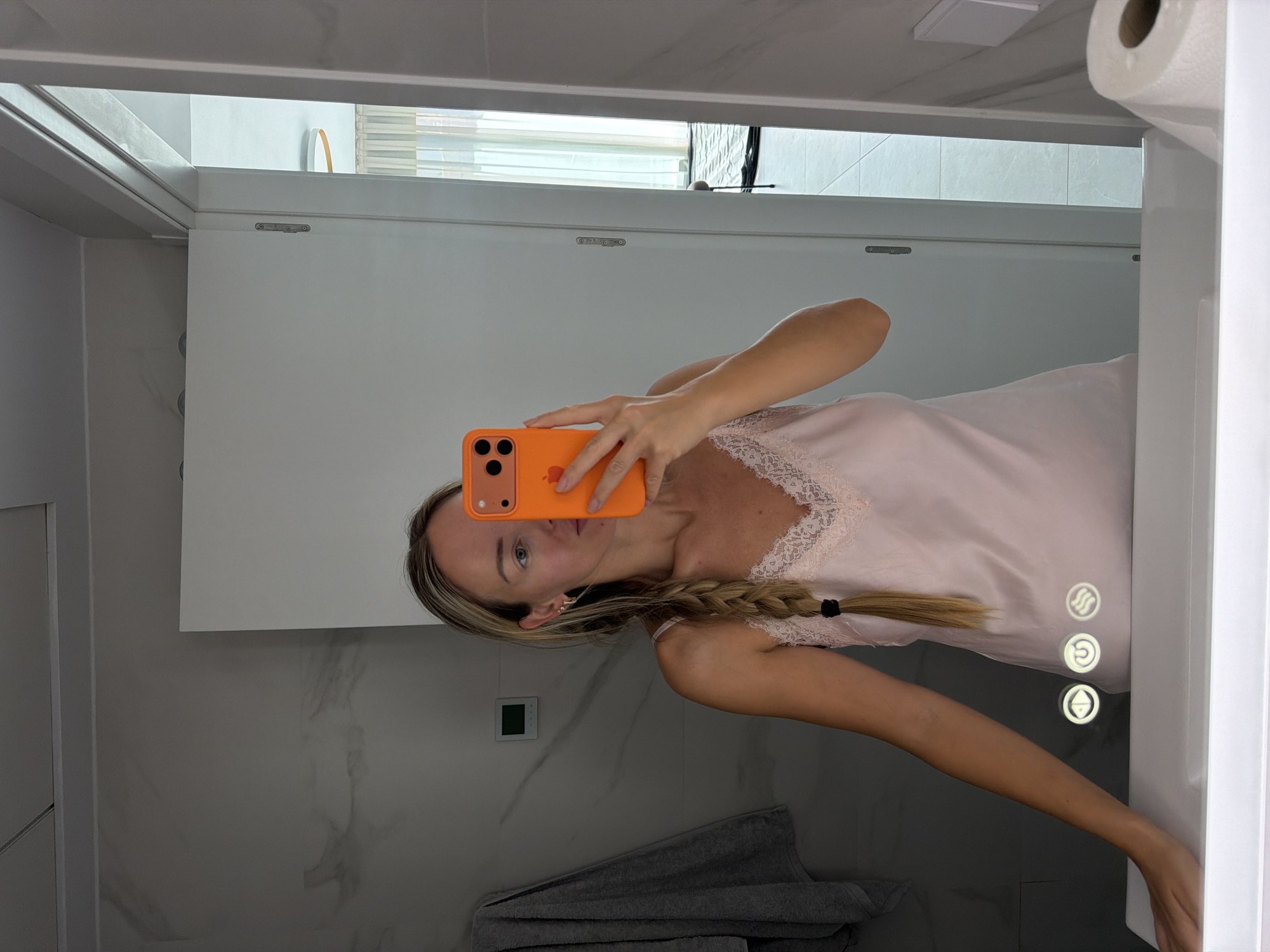 A woman with blonde hair in a braid is taking a mirror selfie in a modern bathroom. She is wearing a light pink slip dress with lace trim. Her left arm is resting on the edge of the white sink. Three illuminated icons are visible on the sink's surface. The bathroom features marble-patterned walls and a white door. In the reflection, a hallway with sheer curtains and a circular light fixture is visible. The scene is set in the city of Finestrat, Spain.