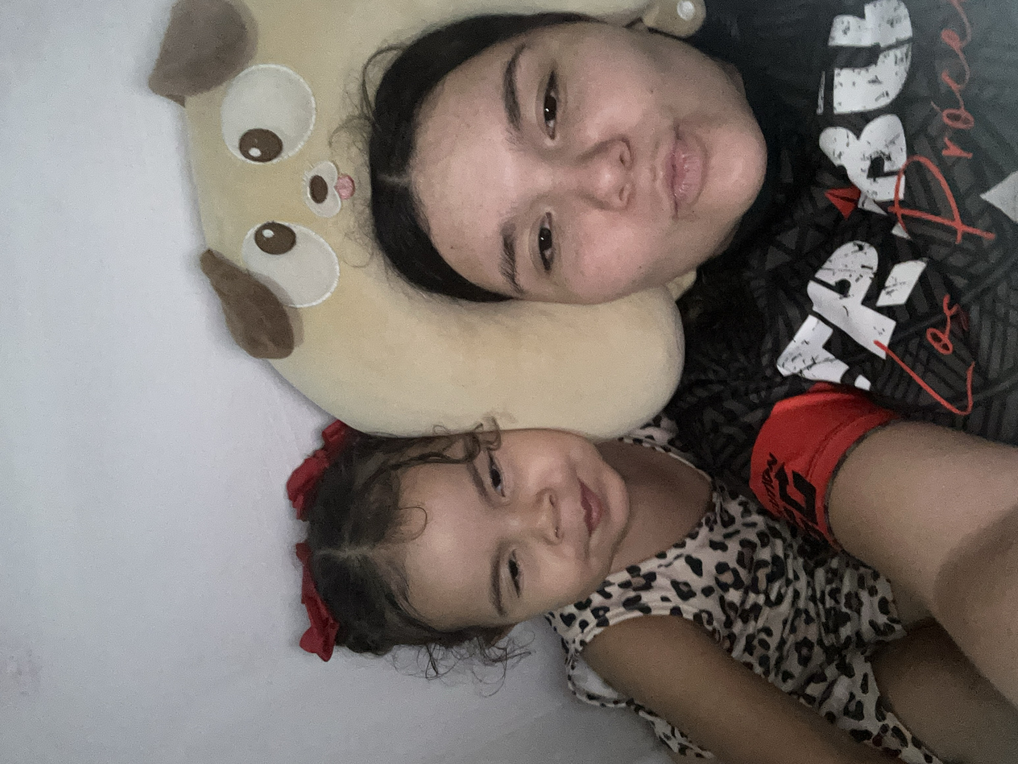 A woman and a young child are lying on a white surface, possibly a bed. The woman is lying on a beige pillow shaped like a dog. The woman has dark hair. She is wearing a dark shirt with white and red lettering and graphics. The child, also with dark hair styled with a red bow, is wearing an animal-print dress. No information allows to know for sure the location is San Juan de Los Morros, Venezuela.