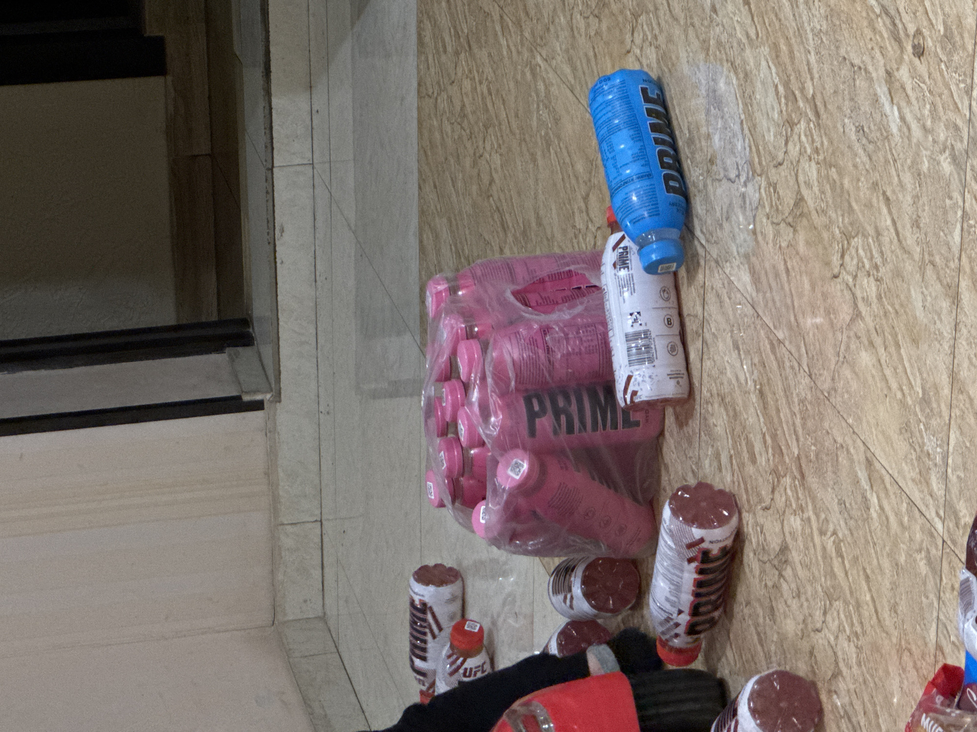 The image depicts a collection of Prime Hydration sports drinks on what appears to be a tiled floor, possibly indoors, given the presence of a staircase in the background. Several bottles are visible, with colors including pink, blue, white, and a red-brown color. One case of pink Prime drinks remains sealed in a plastic bag.
There is no information to identify the exact location in Yerevan, Armenia.