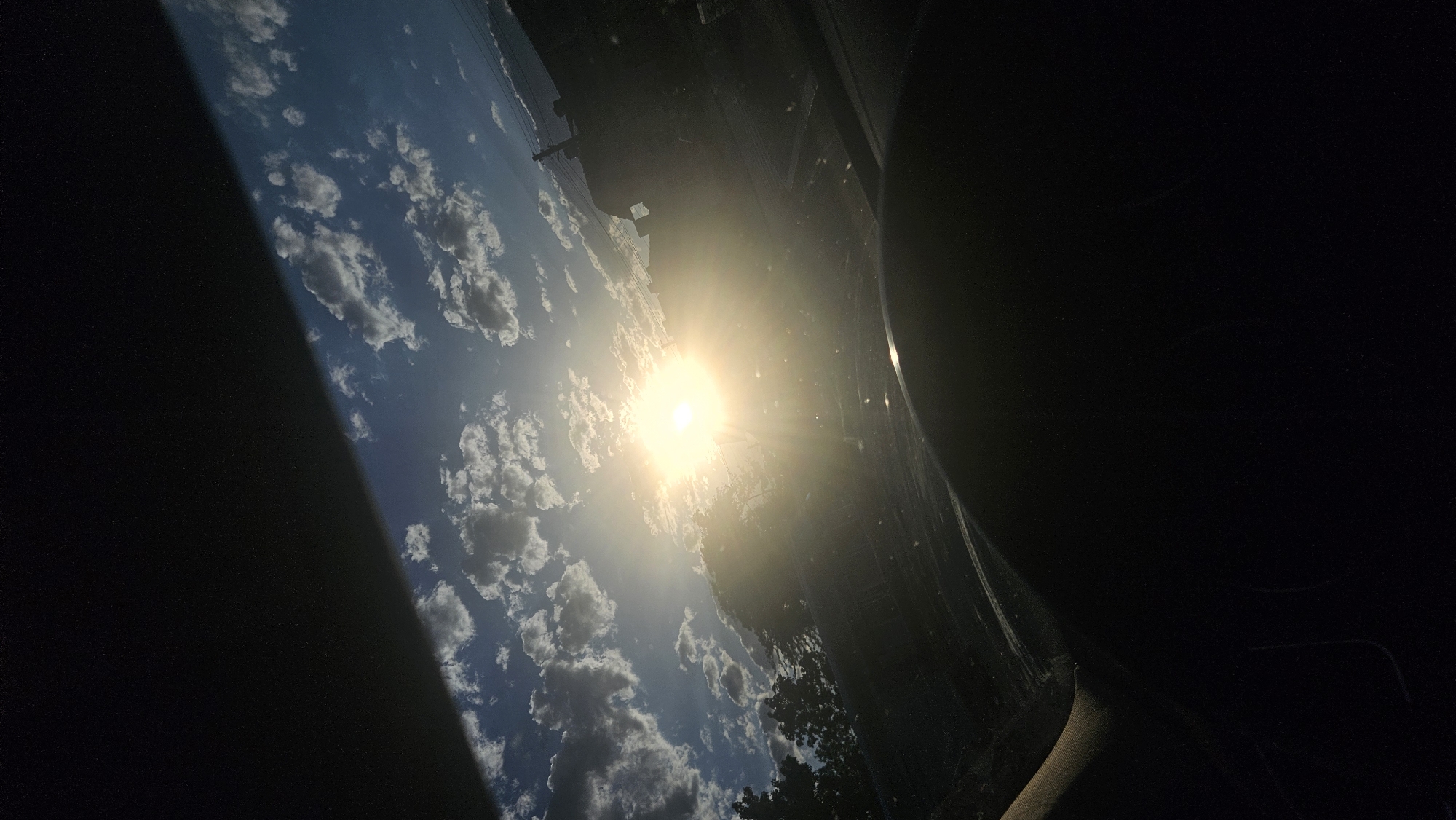 The image shows a sunburst through clouds, viewed from inside a vehicle. The sun is bright and central, with rays extending outwards. Fluffy white clouds are scattered across a blue sky. The silhouette of trees and buildings is visible in the distance, against the bright sky. The foreground is dominated by the dark interior of the car, including what appears to be a steering wheel or dashboard. The overall lighting suggests it is daytime, possibly late afternoon due to the angle of the sun. The location is described as Unknown, Unknown.