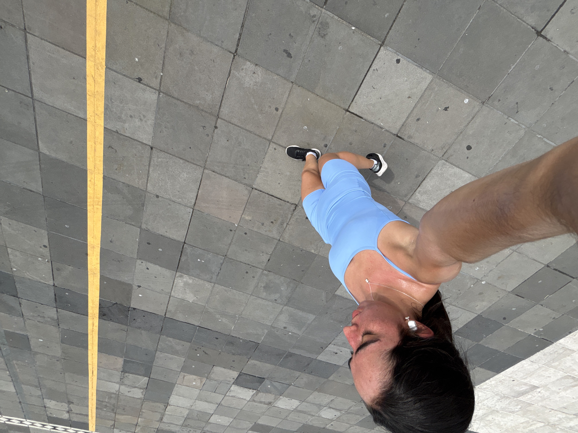 A person wearing a light blue athletic outfit and black and white sneakers stands on a paved surface. A yellow line runs horizontally across the pavement behind the person. The person is looking down and to the right, with their right arm extended and holding what appears to be a smartphone, indicating they are taking a selfie. Their hair is dark and pulled back, and they are wearing white earbuds. The environment suggests an outdoor urban setting, possibly a walkway or plaza. The pavement is composed of large, square-shaped gray tiles arranged in a diagonal pattern. The lighting indicates it is daytime. The image quality is clear and shows good detail of the subject and surroundings. There are no discernible landmarks that would pinpoint the exact location within Veracruz City, Mexico.