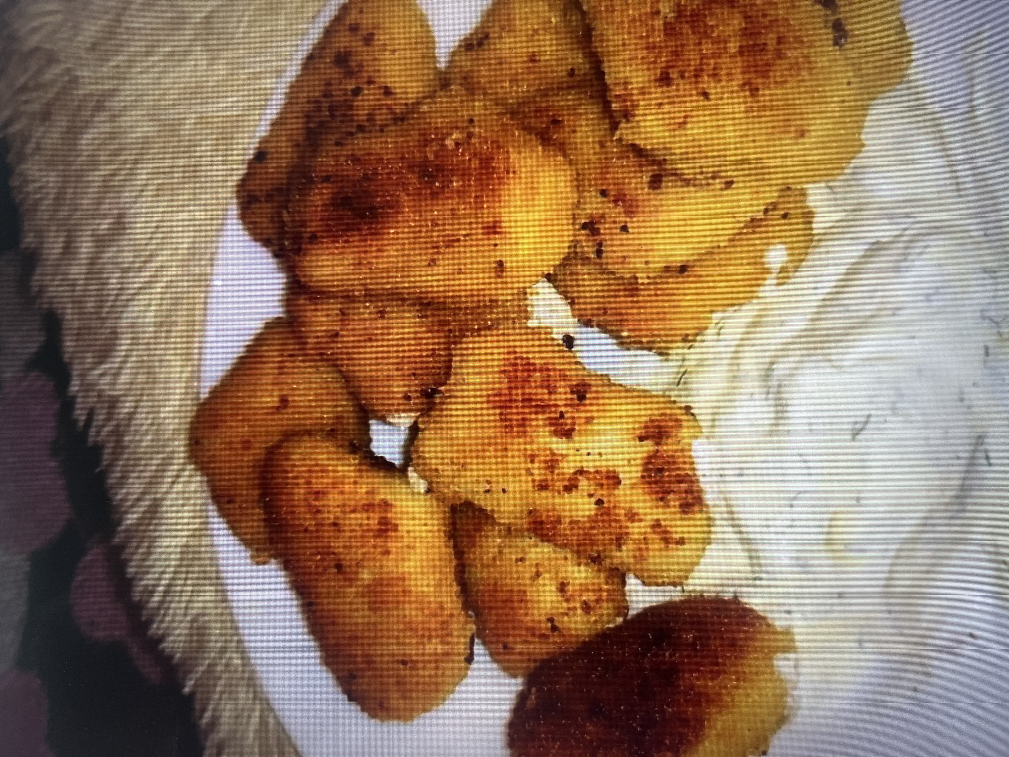 A plate of golden-brown fried food, possibly chicken tenders or fish sticks, is arranged on a white plate. The fried items are coated in a crispy breading and some pieces show darker, more browned areas indicating a good crisp. A white, creamy dipping sauce with visible green flecks, likely herbs such as dill or parsley, is spread across the bottom of the plate. The plate is set on a soft, fuzzy surface that appears to be a blanket or rug, with a blurred background of dark and purplish colors, suggesting an indoor setting. There are no people or specific landmarks visible to determine a location in Moscow, Russia.