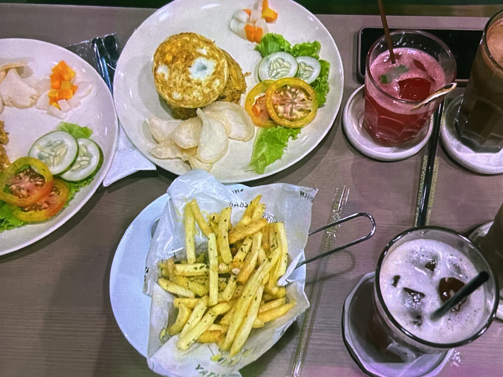 The image shows a top-down view of a table at a restaurant in Cadek, Indonesia. Two plates of food and two drinks are visible. One plate contains french fries served in a paper liner, with tongs to pick them up. The other plate features a fried egg, crackers, and a salad of lettuce, tomatoes, and cucumbers. Next to the plates are two glasses of drinks, one with ice and a straw, and the other a pinkish beverage with ice and a straw. The table surface is a dark, textured material, possibly wood or laminate. The lighting is dim, suggesting an indoor setting. There are no people visible in the image.