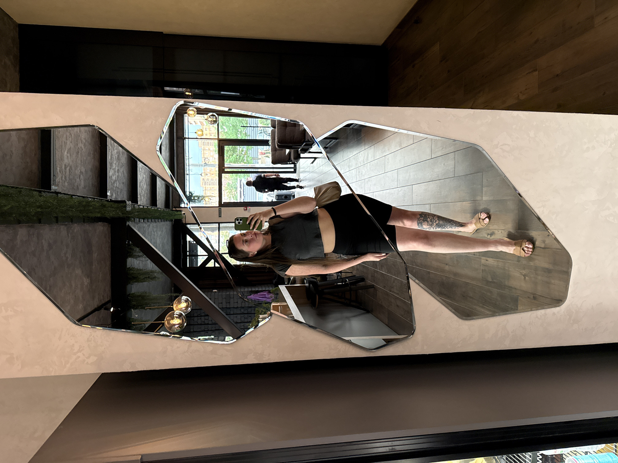 A woman with long brown hair is taking a mirror selfie in what appears to be a cafe or restaurant. She is wearing a dark grey crop top, black shorts, and tan slides. She is holding a smartphone in her left hand, and a light tan purse is slung over her right shoulder. The mirror is irregularly shaped and reflects the interior of the establishment, including exposed beams, hanging plants, and industrial-style lighting. In the background of the mirror's reflection, a man in a dark shirt and pants is walking out of a set of glass doors. The floor in the reflection is tiled, and further beyond the doors, there is an outdoor area with buildings visible. The woman has a tattoo on her left calf.