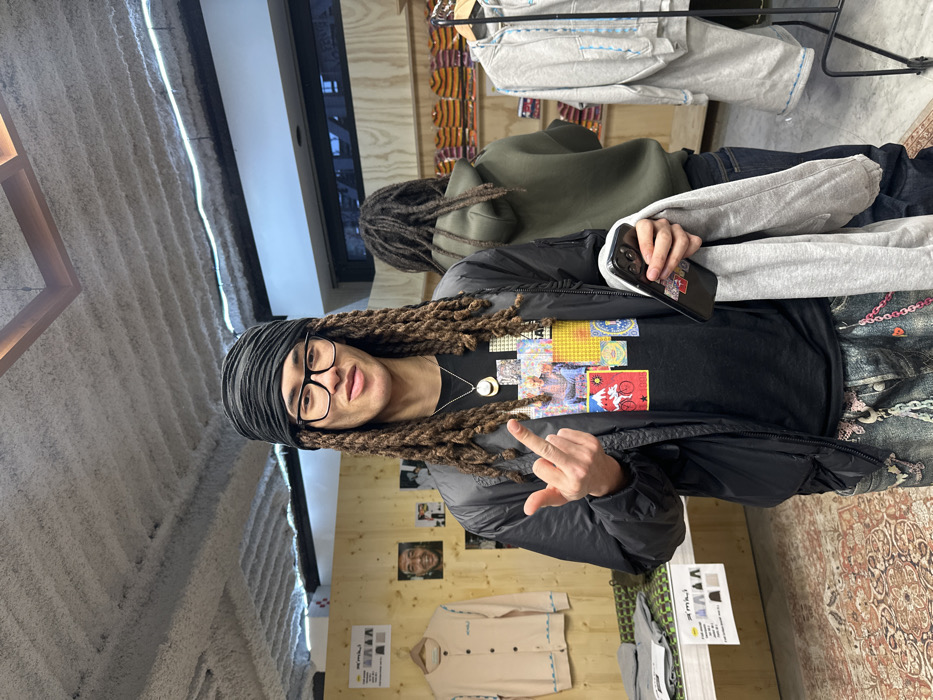 An indoor retail space in Paris, France, features a young adult male with dreadlocks and black-framed glasses, wearing a black headwrap, centrally positioned and facing the viewer. He wears a black bomber jacket over a black t-shirt with a colorful, collage-style graphic print, and distressed light-wash denim jeans adorned with multiple pink and silver chains. His right hand is raised with the middle finger extended, while his left hand holds a black smartphone and a folded grey garment. A silver necklace with a circular pendant is visible.
In the background, a second individual with dreadlocks stands facing away from the camera, wearing an olive green hooded sweatshirt and dark jeans. The retail environment includes light wooden display panels on the left, showcasing a cream-colored cardigan and framed images, one depicting a smiling man. Below the cardigan, a table displays folded grey clothing and an information card partially visible as "s'miki". Behind the second person, wooden shelves hold stacks of neatly folded, striped garments, primarily in orange and cream tones. To the right, a black metal clothing rack displays grey hooded sweatshirts with light blue stitching.
The floor in the foreground is covered by a patterned rug in shades of red, beige, and brown, transitioning to lighter-colored tiles further back. The ceiling is exposed concrete with visible beams and textures. A horizontal white neon light strip runs along the wall-ceiling junction in the background, and a square light fixture is suspended from the ceiling. A large window in the background reflects exterior urban buildings.