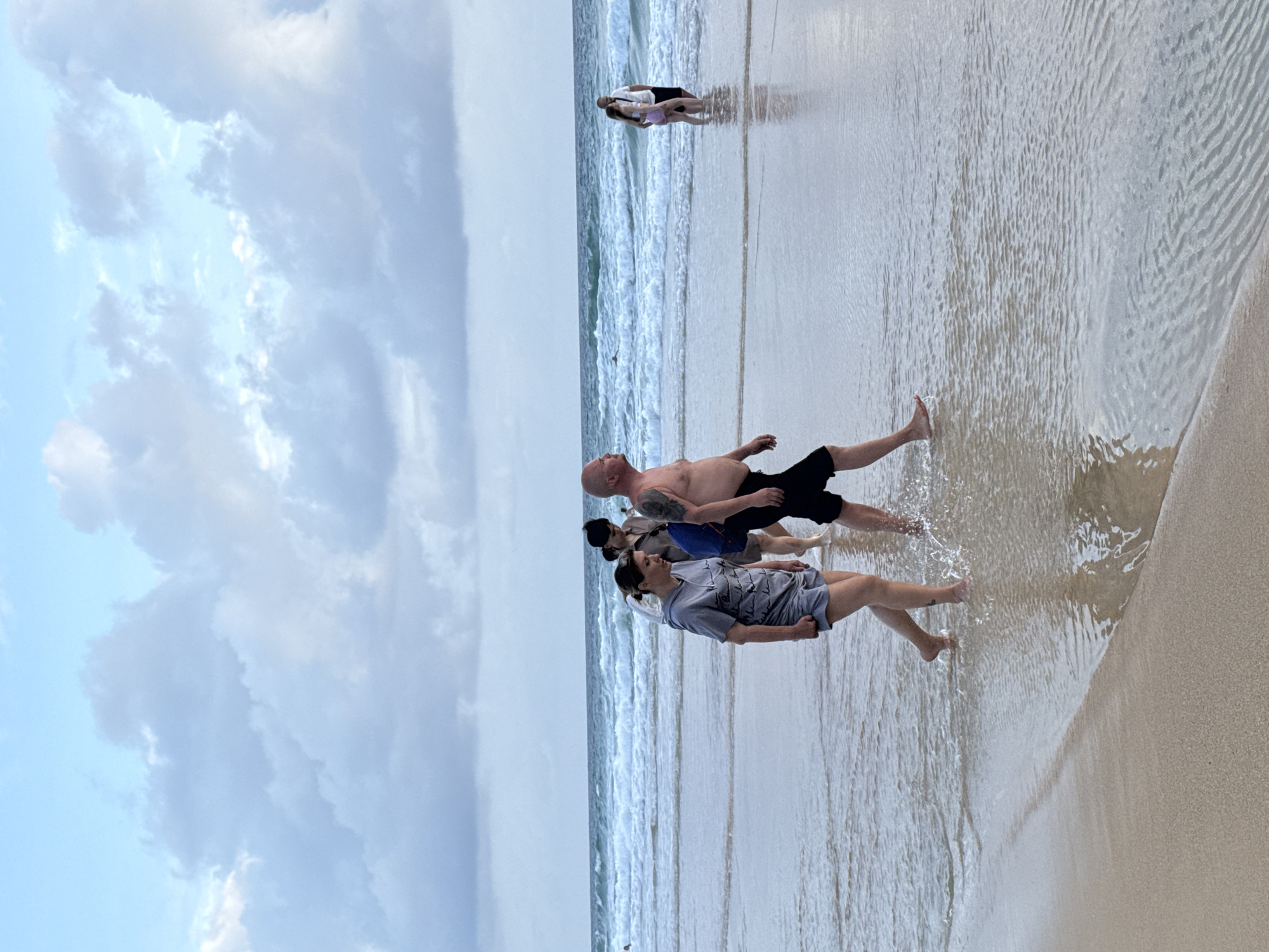 In Karon, Thailand, several people are walking on a beach. A man and a woman in the foreground are walking through shallow water. The man is shirtless and wearing black shorts, while the woman is wearing a grey t-shirt and grey shorts. Behind them, a group of four people walks further up the beach, with two of them carrying a blue bag. In the distance, a couple stands in the water near the shore, with the woman wearing a pink and white swimsuit and the man wearing a white t-shirt and black shorts. The ocean has moderate waves, and the sky is overcast with scattered clouds. The sand is wet and reflects the light.
